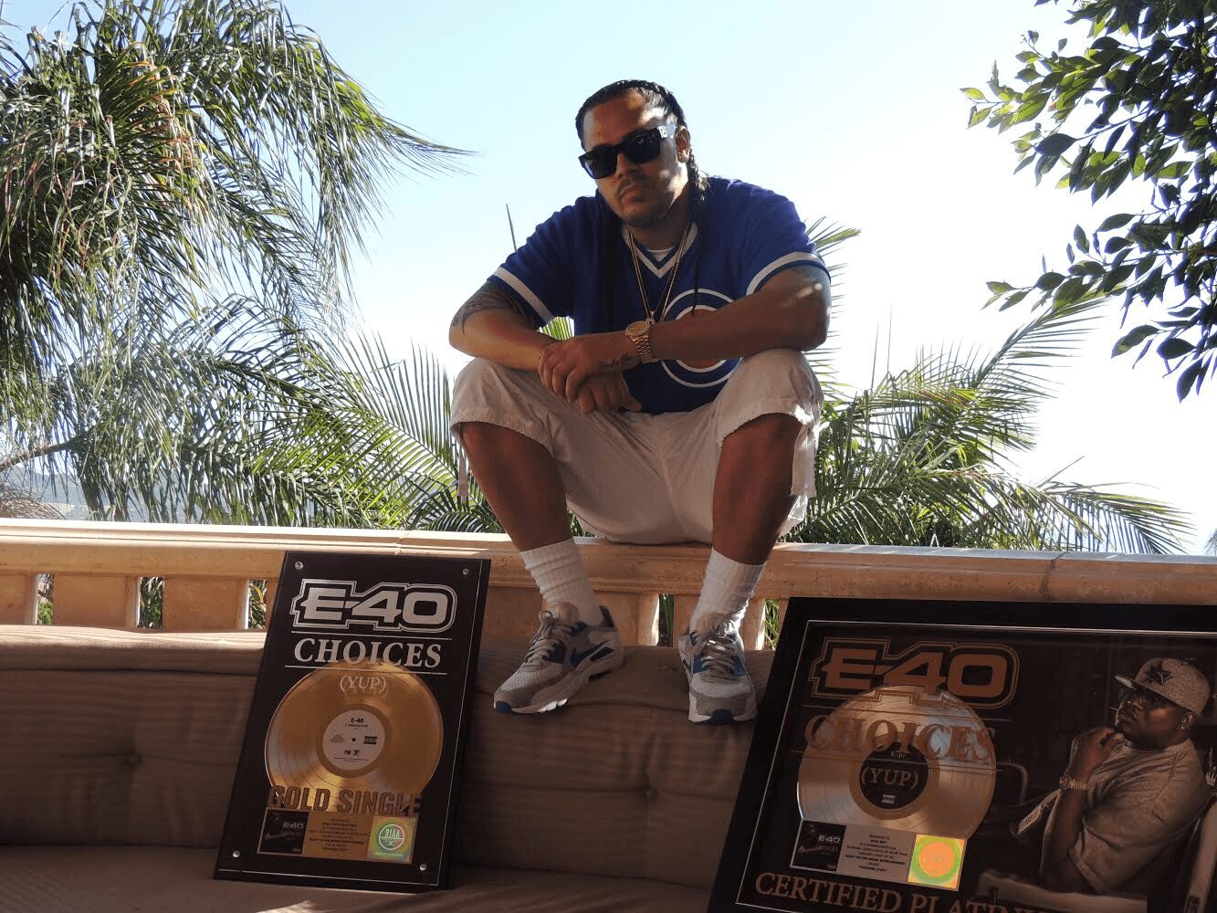 Person crouching on a ledge outdoors with palm trees in background, wearing sunglasses, dark shirt, and light shorts.