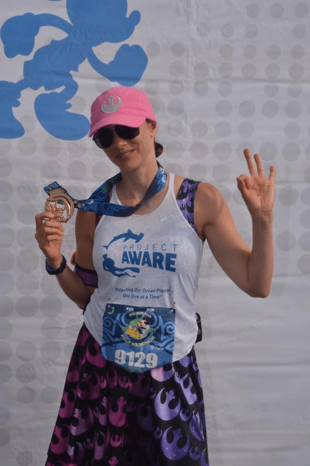Person wearing pink cap, sunglasses, holding a medal, making a peace sign, in athletic outfit with bib number 9129.