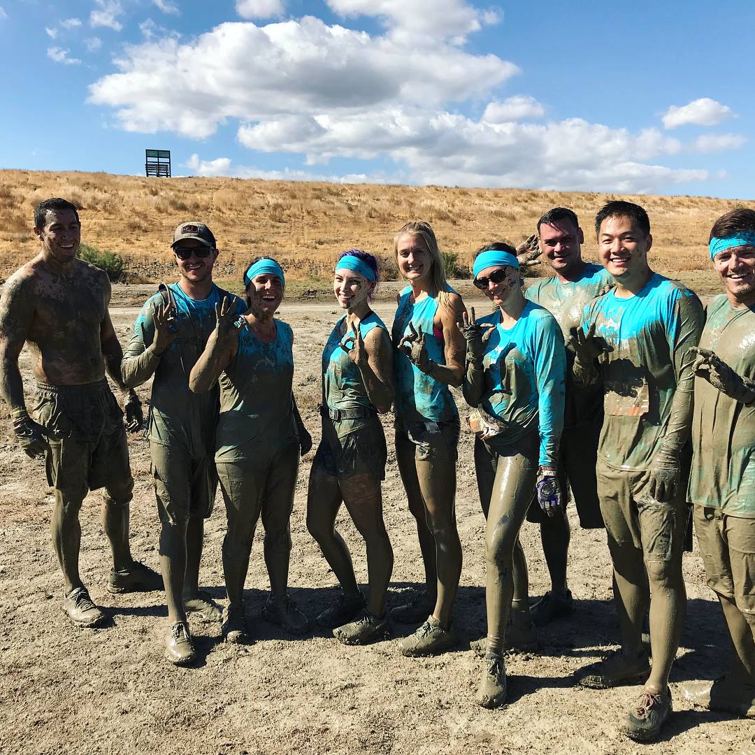 Group of nine people standing outdoors on a dirt path, some with blue headbands, in sunny weather.
