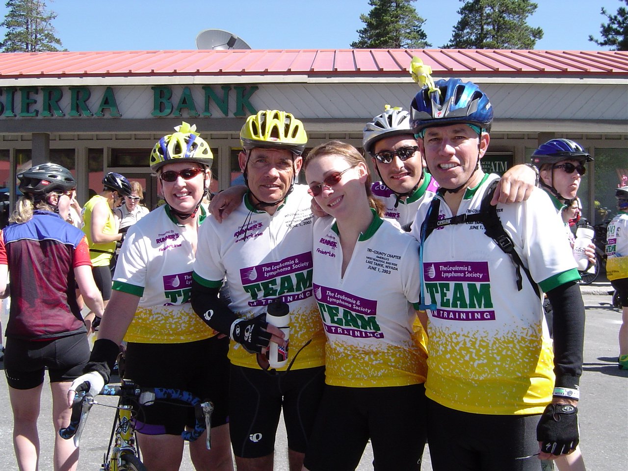 Group of six cyclists in helmets and cycling gear standing together outdoors, smiling, in front of a bank building.