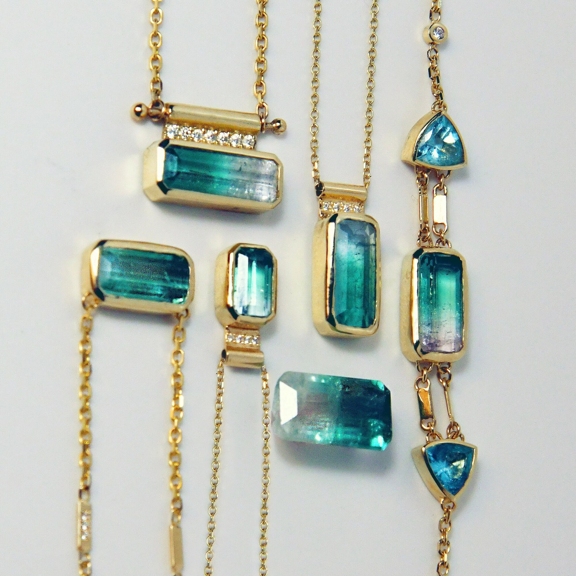 Collection of gold jewelry with blue-green rectangular and triangular gemstones, including necklaces and bracelets, on a white background.