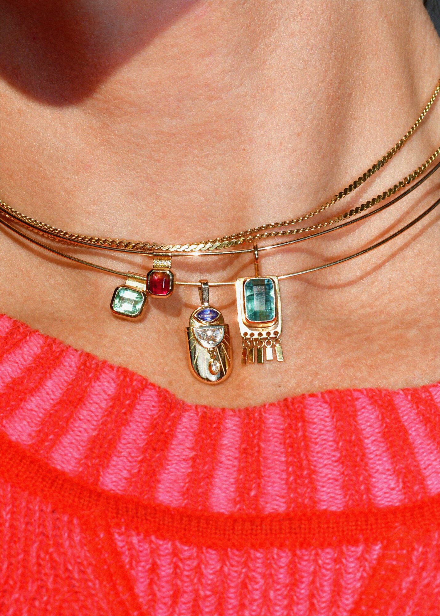 Close-up of a person's neck with layered gold necklaces and colorful pendants, wearing a pink and red striped top.
