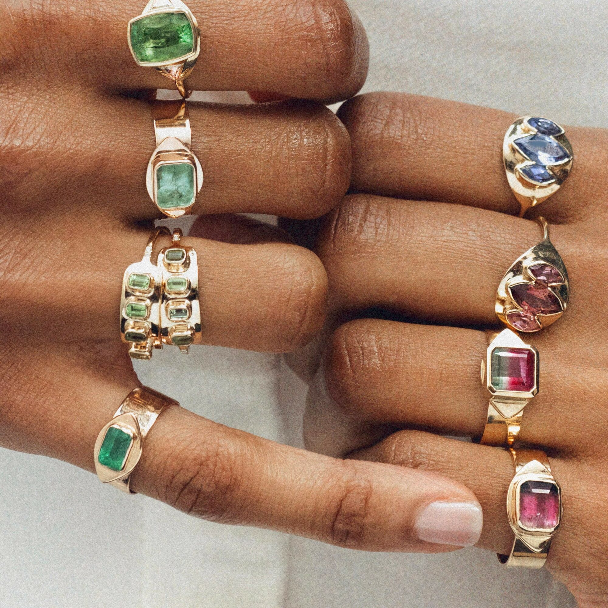 Person's hands with multiple rings featuring colorful gemstones, some with gold bands, on fingers and thumb.