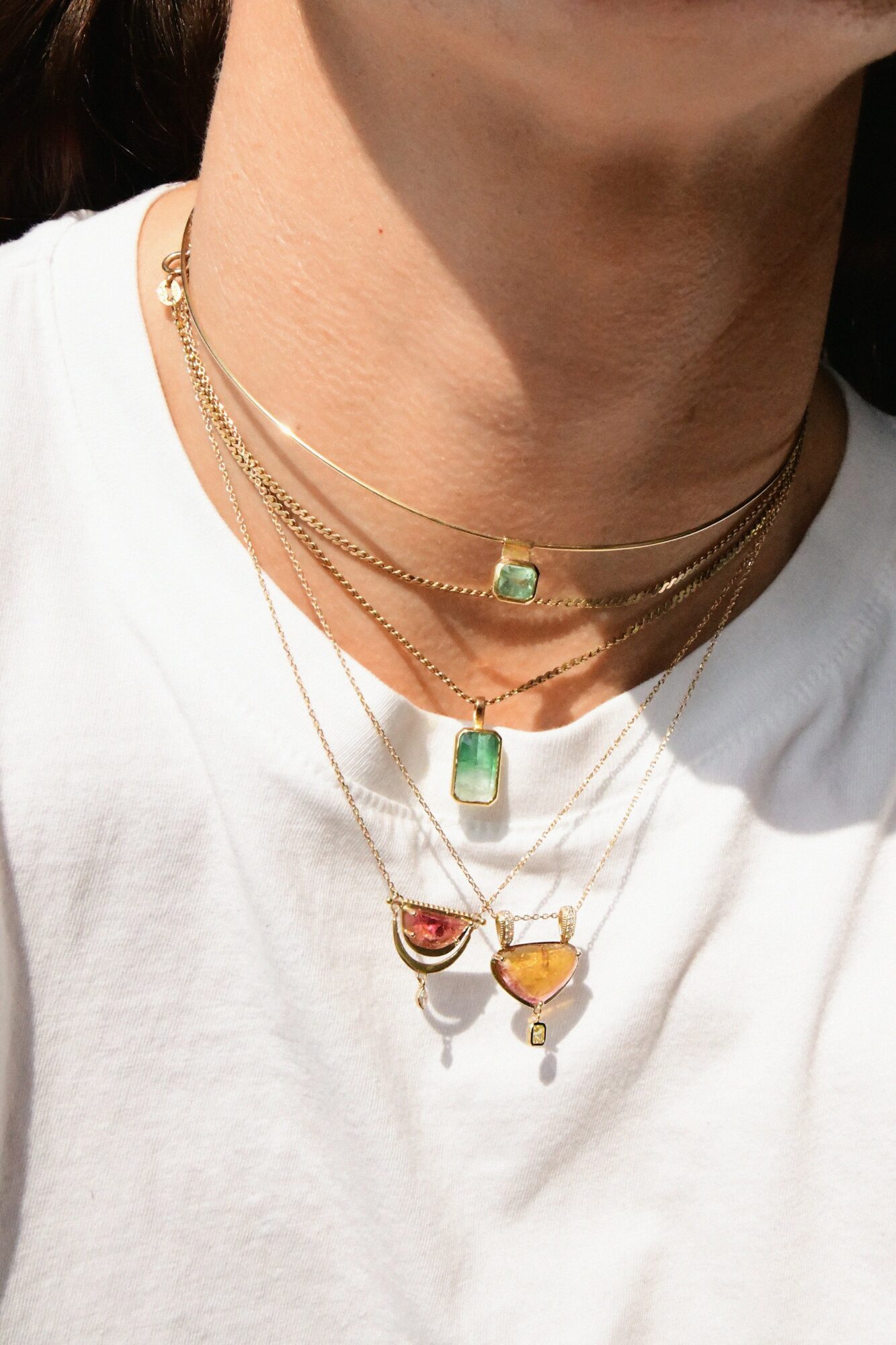 Person wearing layered gold necklaces with colorful gemstone pendants on white shirt.