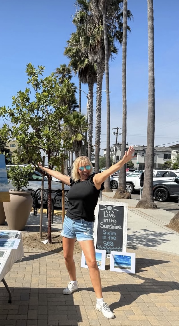 Person with arms raised standing on sidewalk near palm trees and a sign, sunny day with parked cars in background.