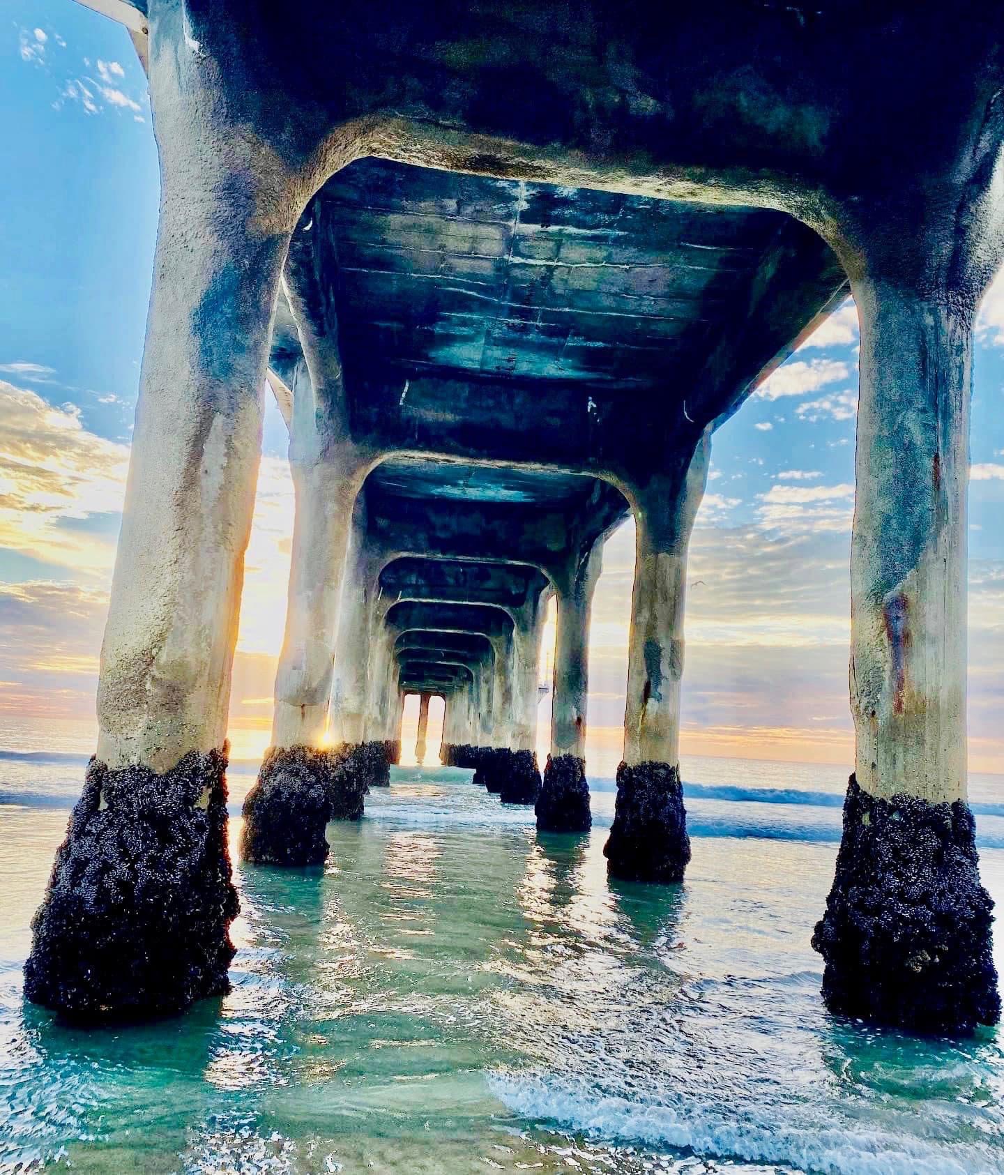 Underwater view of a pier with multiple supports extending into the water, sunlight visible through gaps, sky with clouds.