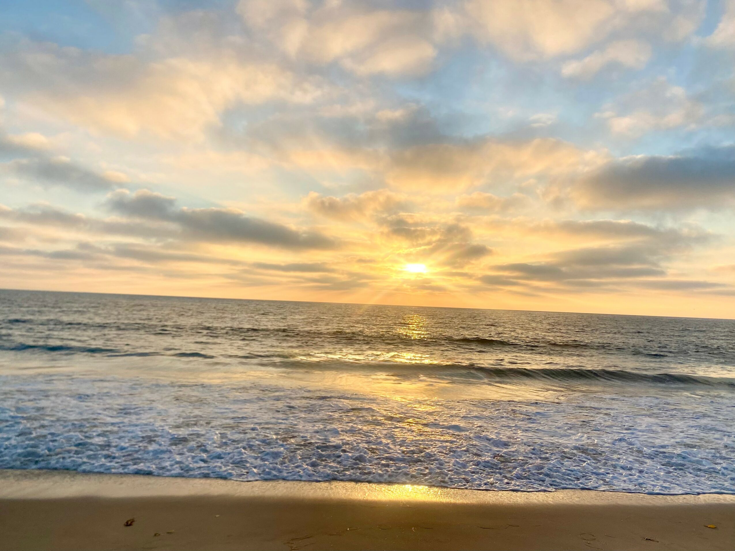Sunset over the ocean with clouds and waves, sandy beach in foreground.