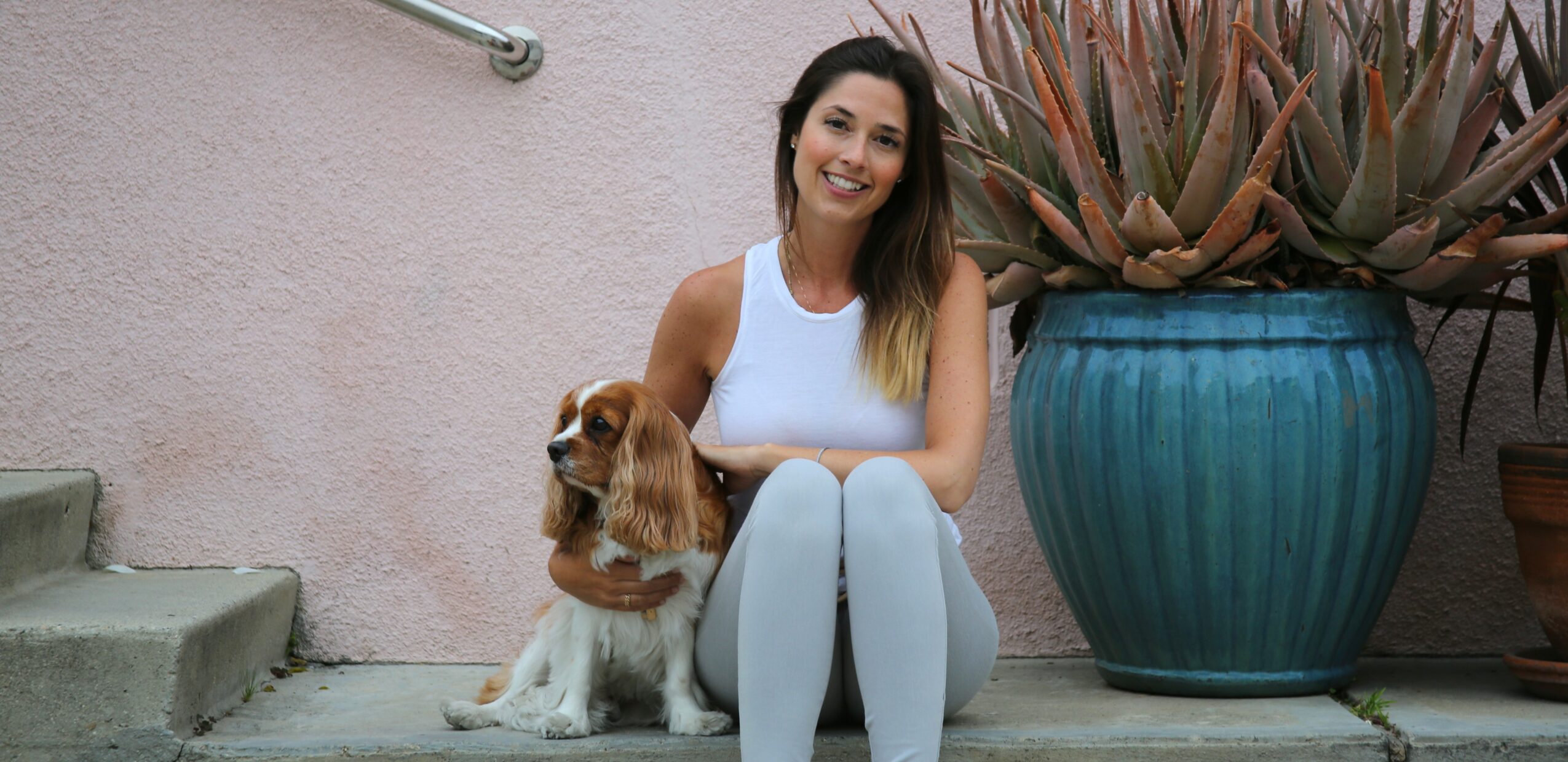 Woman sitting on steps with a dog, large potted plant nearby, smiling outdoors.