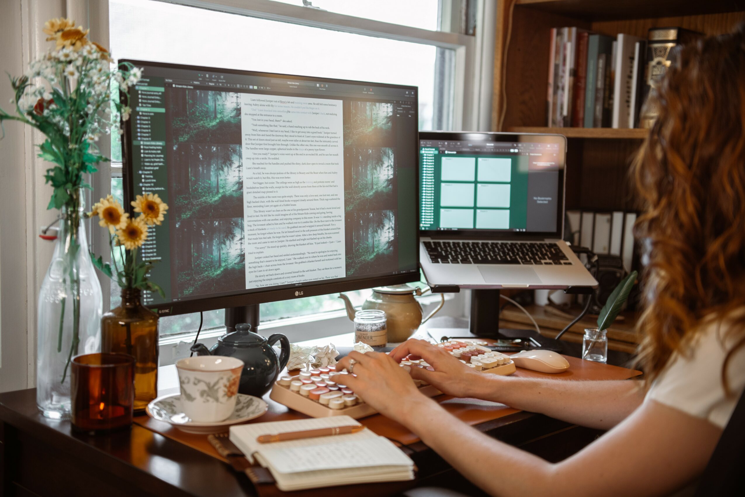Person working at a desk with two monitors, a keyboard, and a notebook, in a room with bookshelves and a window.