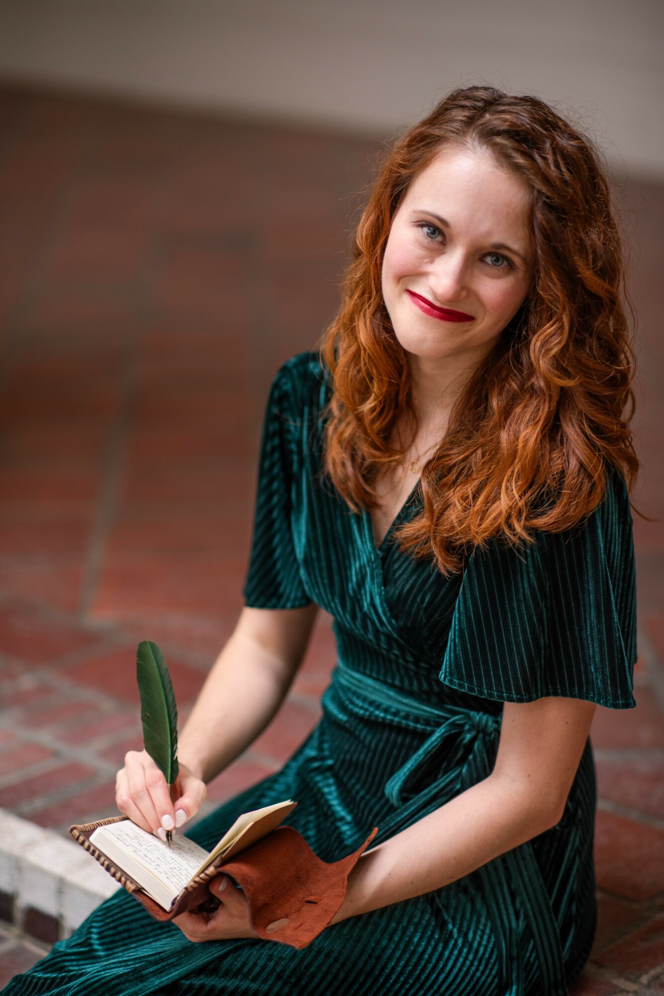 Young woman with curly red hair sitting on a brick surface, smiling, holding a pen and notebook.