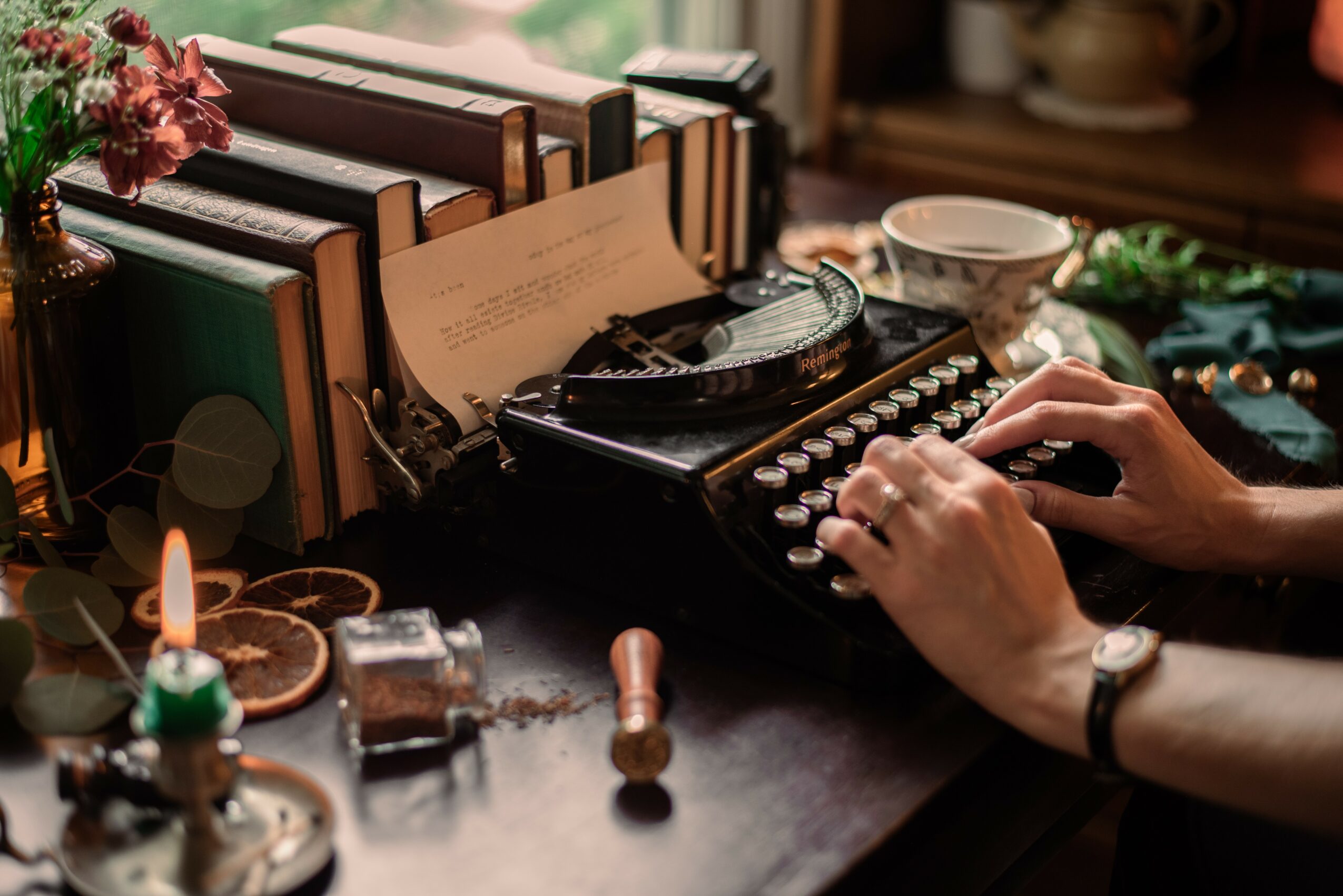 Person typing on a vintage typewriter surrounded by books, candles, and decorative items on a dark wooden table.