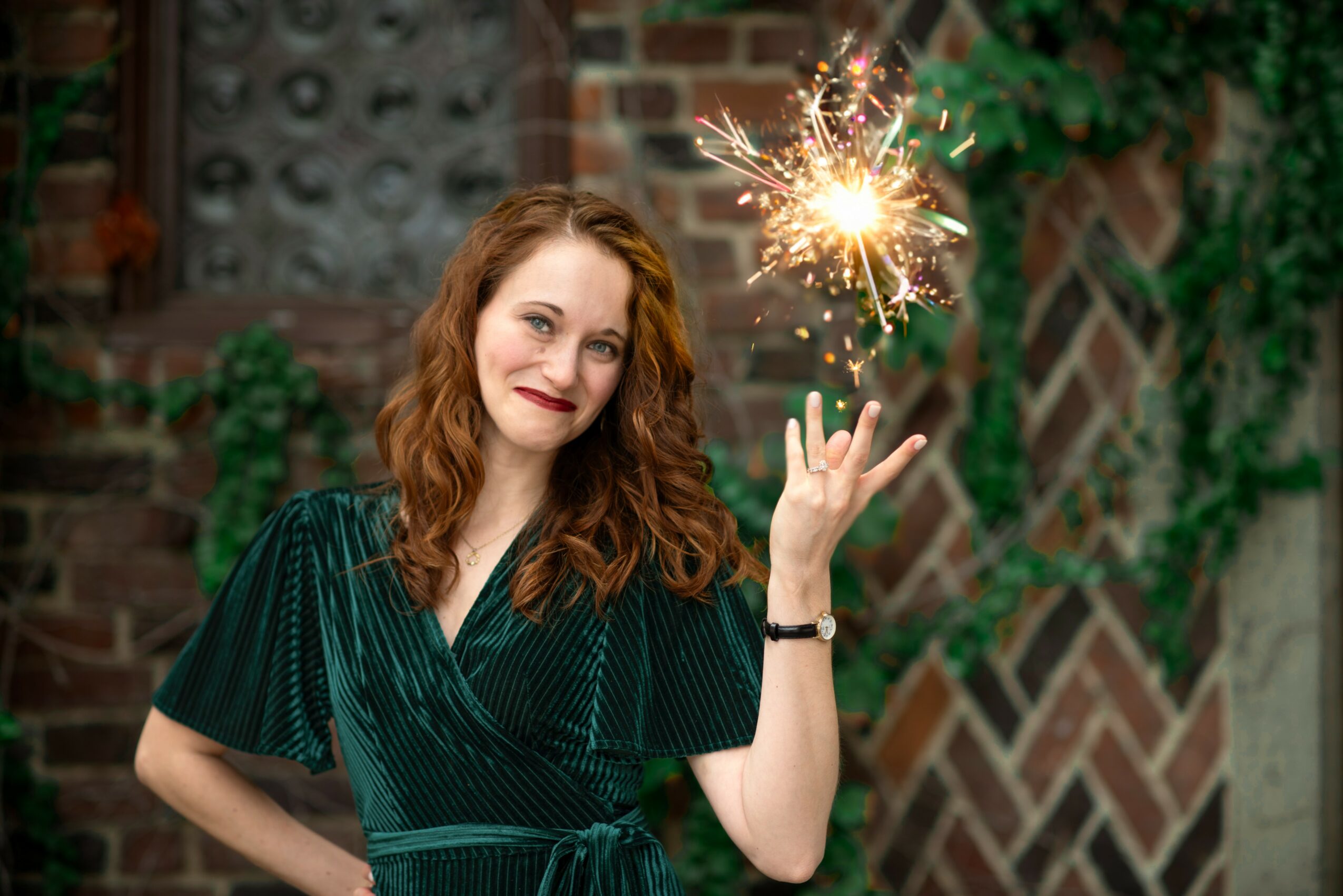 Woman with wavy hair holding a lit sparkler, smiling, in front of a brick wall with greenery.