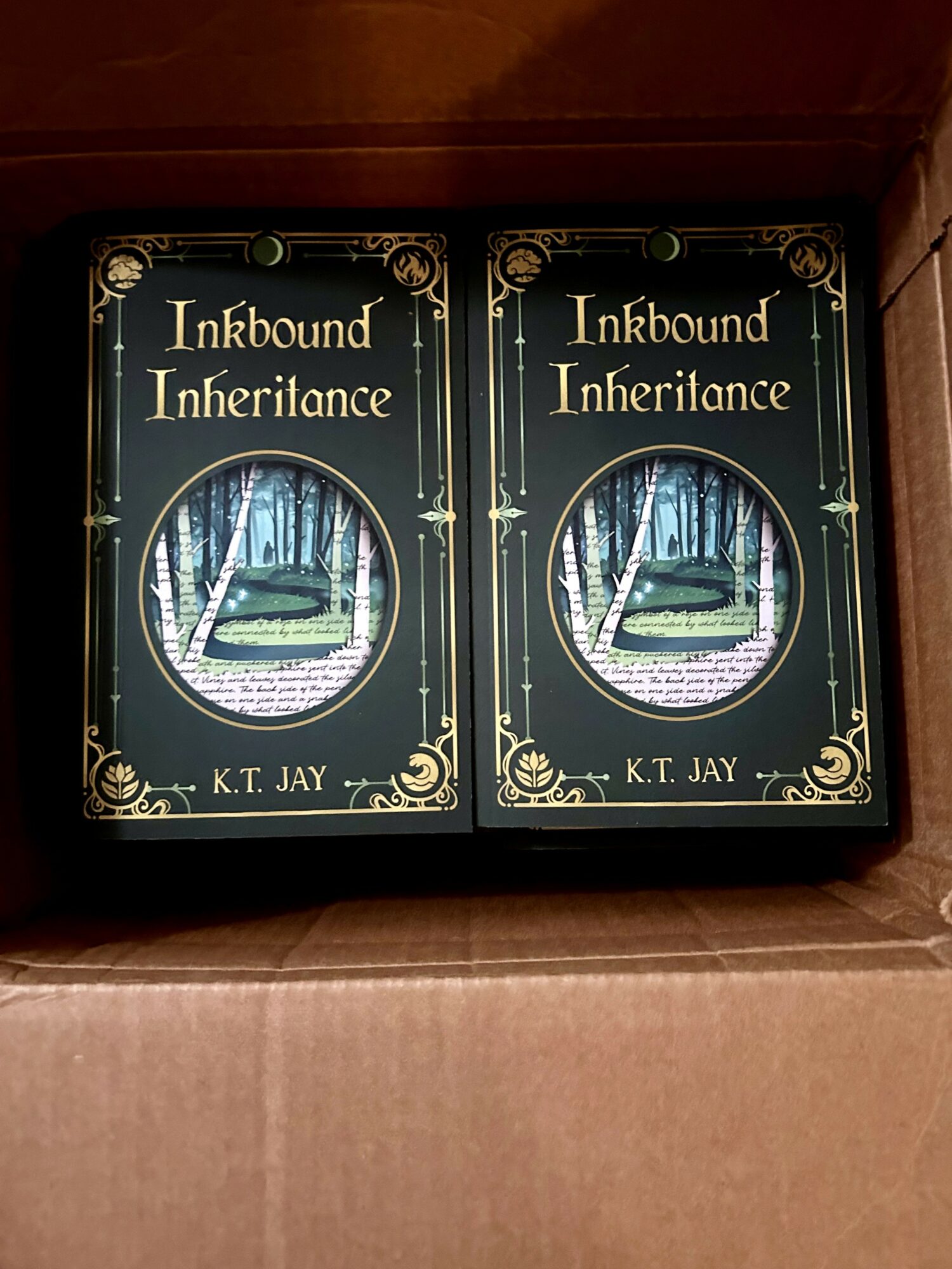 Two copies of the book titled 'Inkbond Inheritance' by K.T. Jay, with a forest scene on the cover, inside a cardboard box.