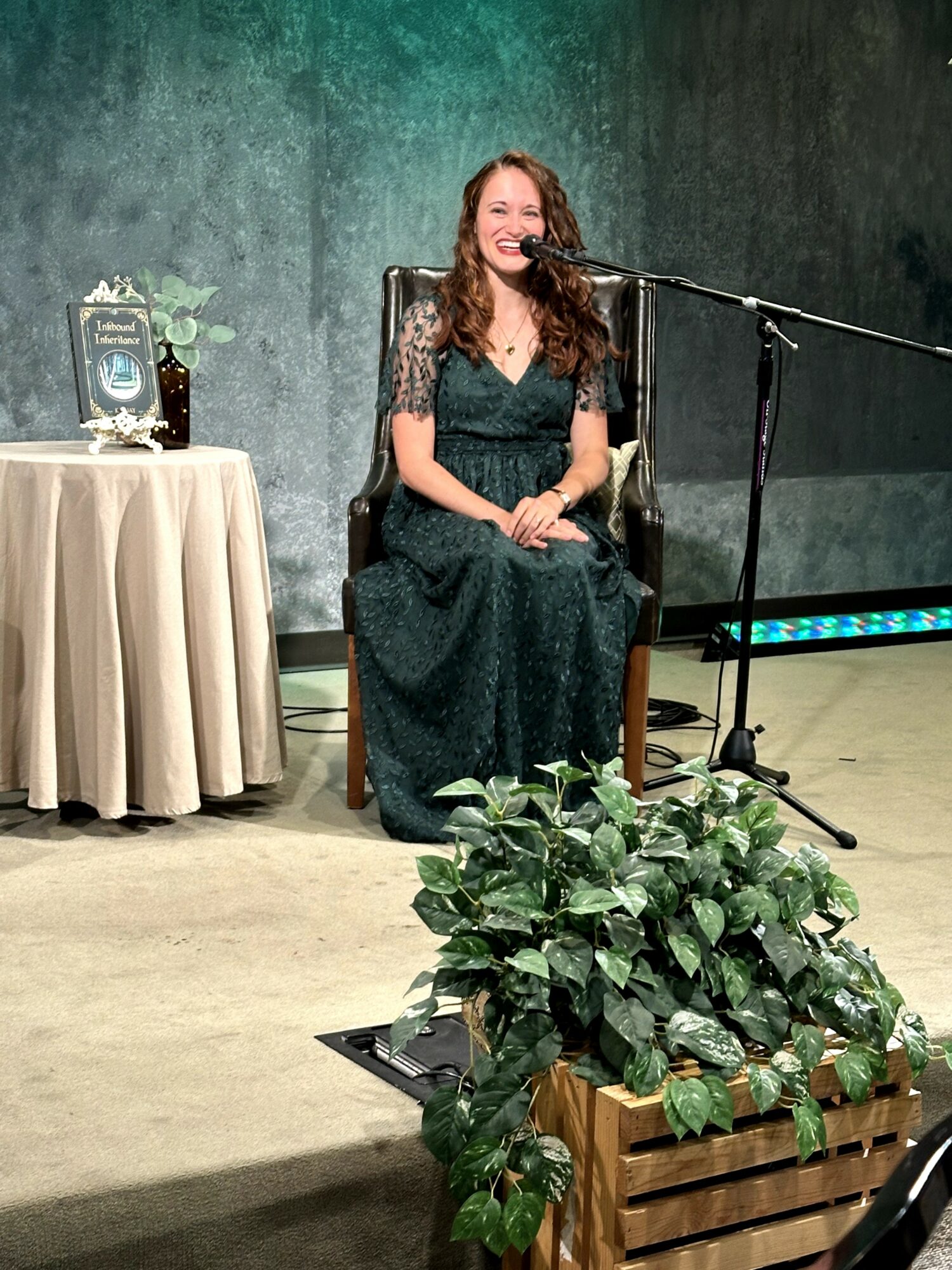 Woman in a dark green dress sitting on a chair, smiling, with a microphone in front, next to a table with a framed photo and flowers.