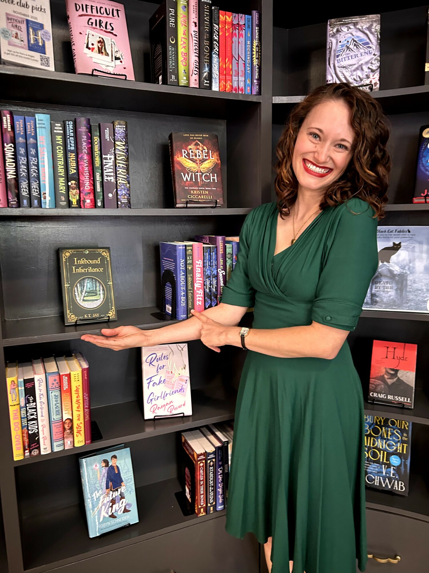 Woman in green dress smiling and pointing at a book on a bookshelf filled with colorful books.