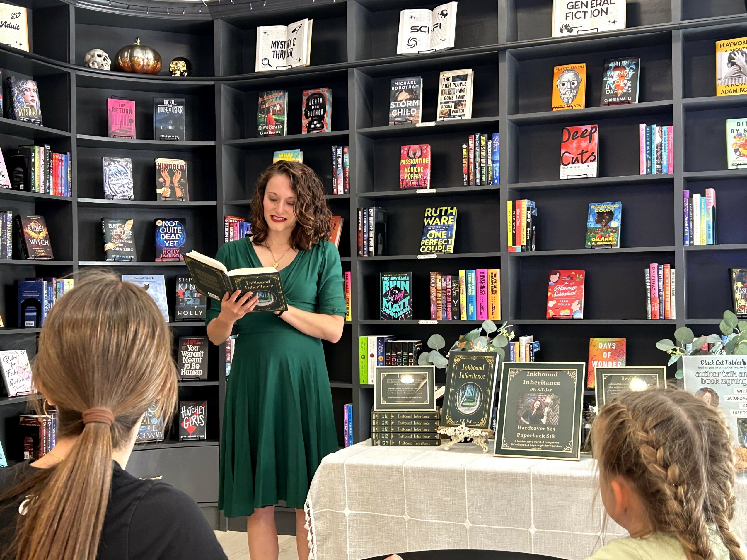 Woman in green dress reading a book to children in a bookstore with shelves of books behind her.