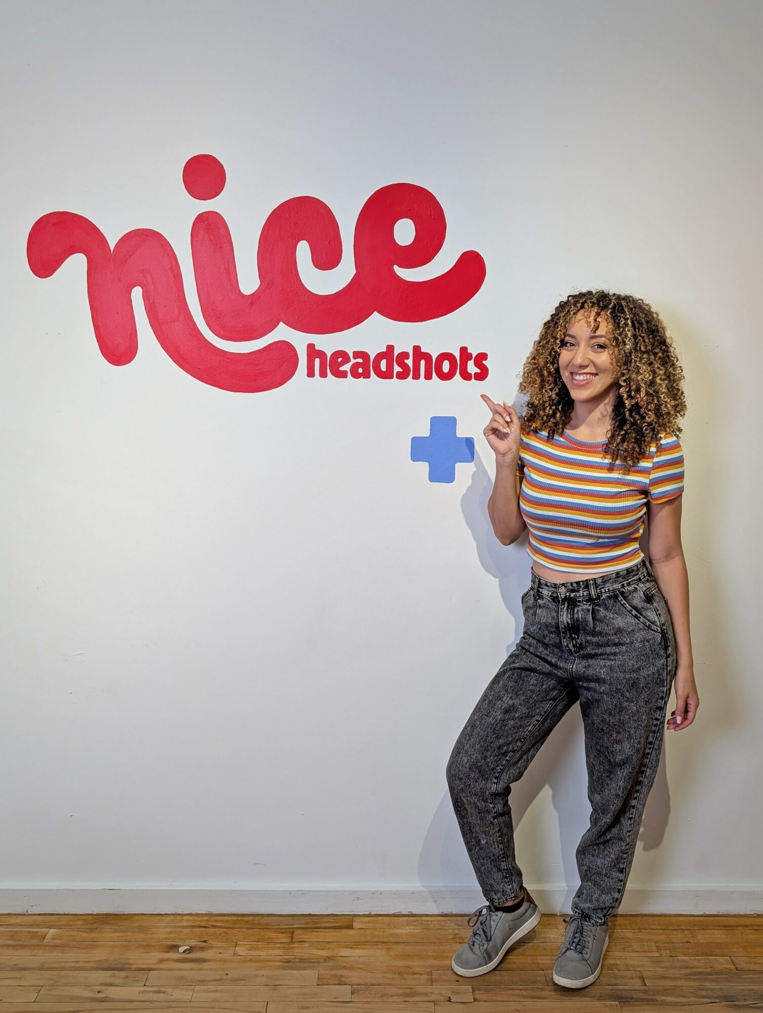 Young woman with curly hair smiling and pointing at a wall with 'nice headshots' logo and a blue plus sign.