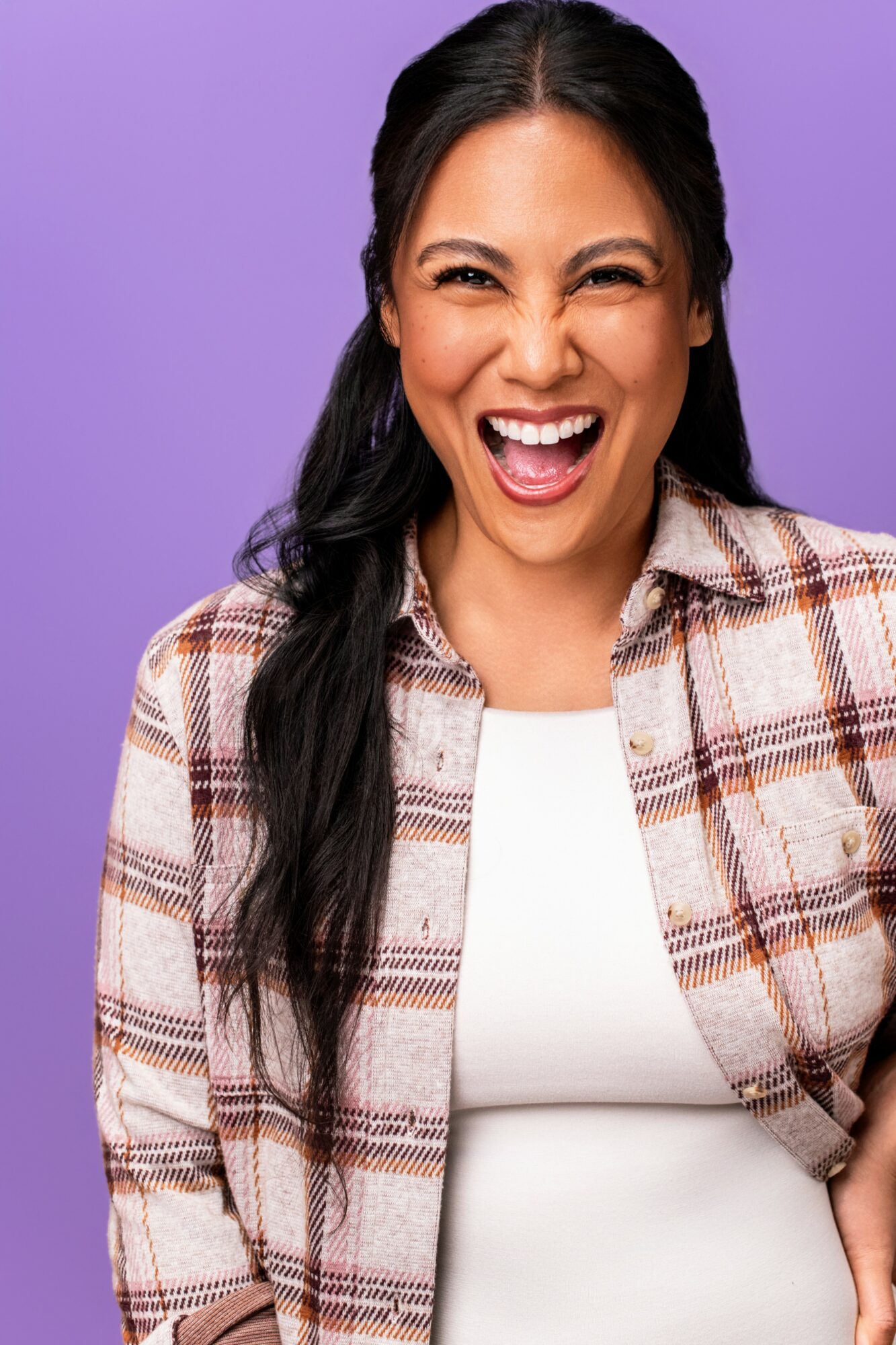 Woman with long dark hair smiling and laughing, wearing a plaid shirt and white top, against a purple background.