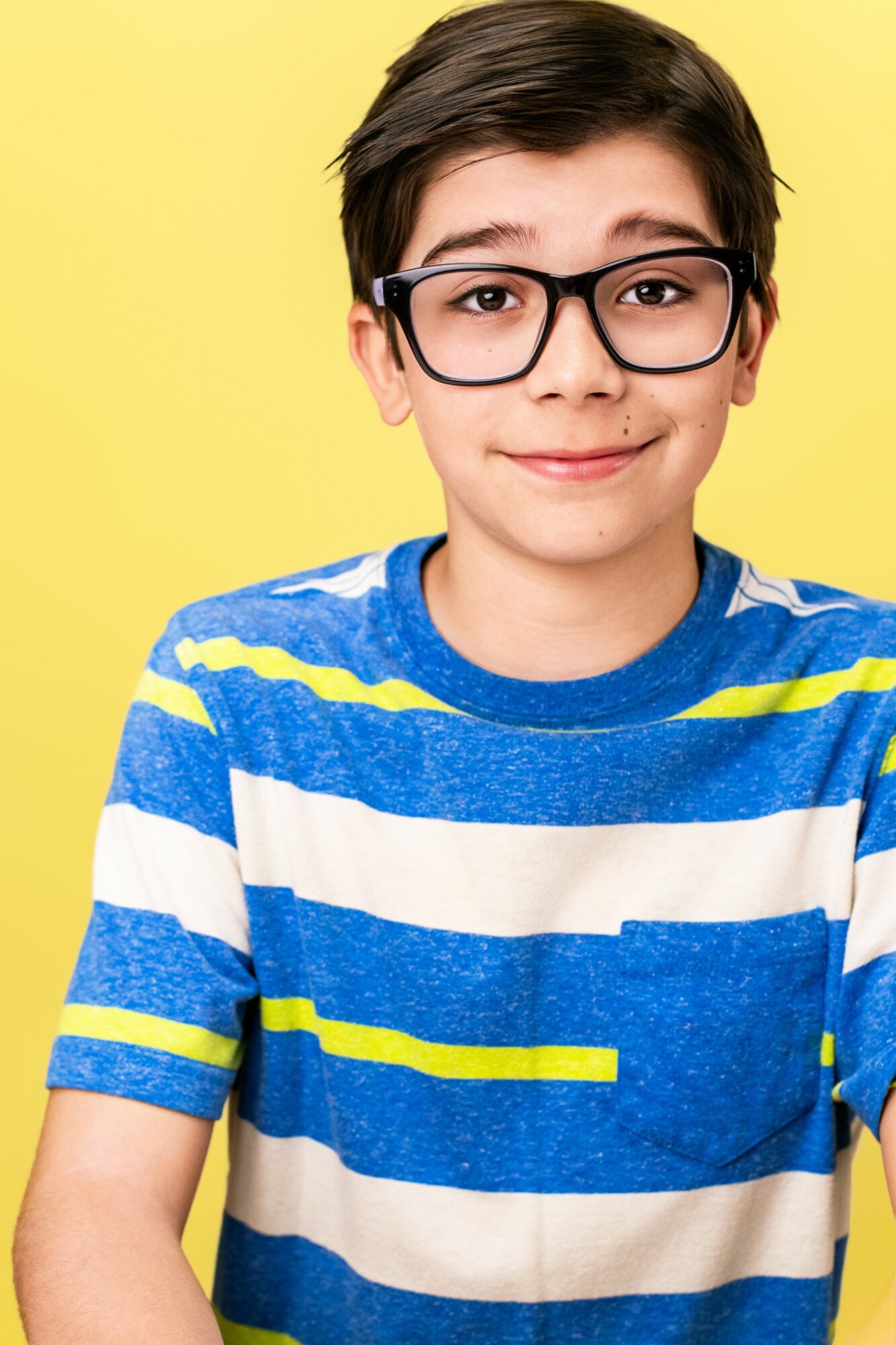 Boy with glasses wearing a blue and white striped shirt, smiling, yellow background.