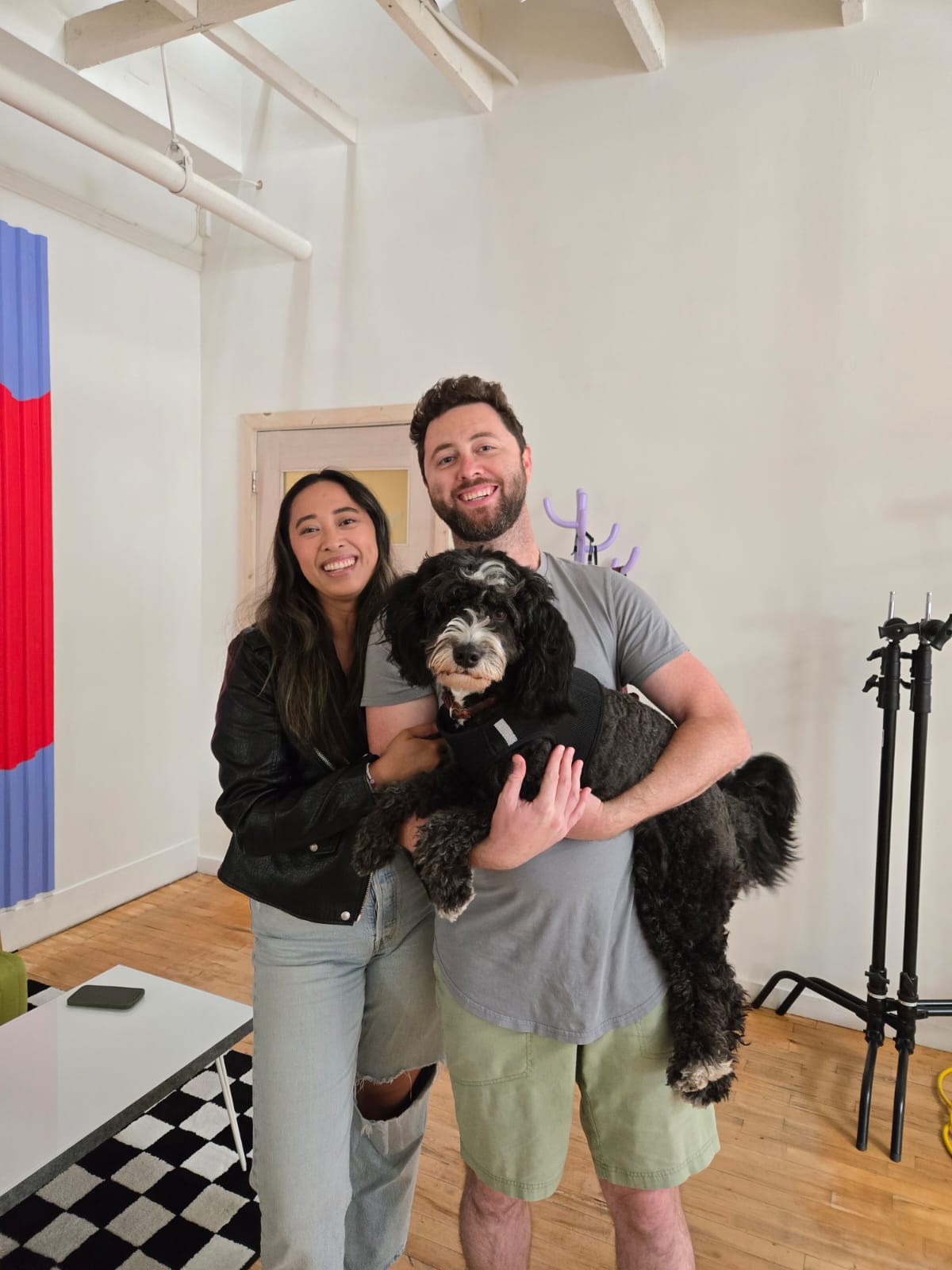 Two people smiling and holding a large black and white dog indoors, with a white wall and wooden floor.