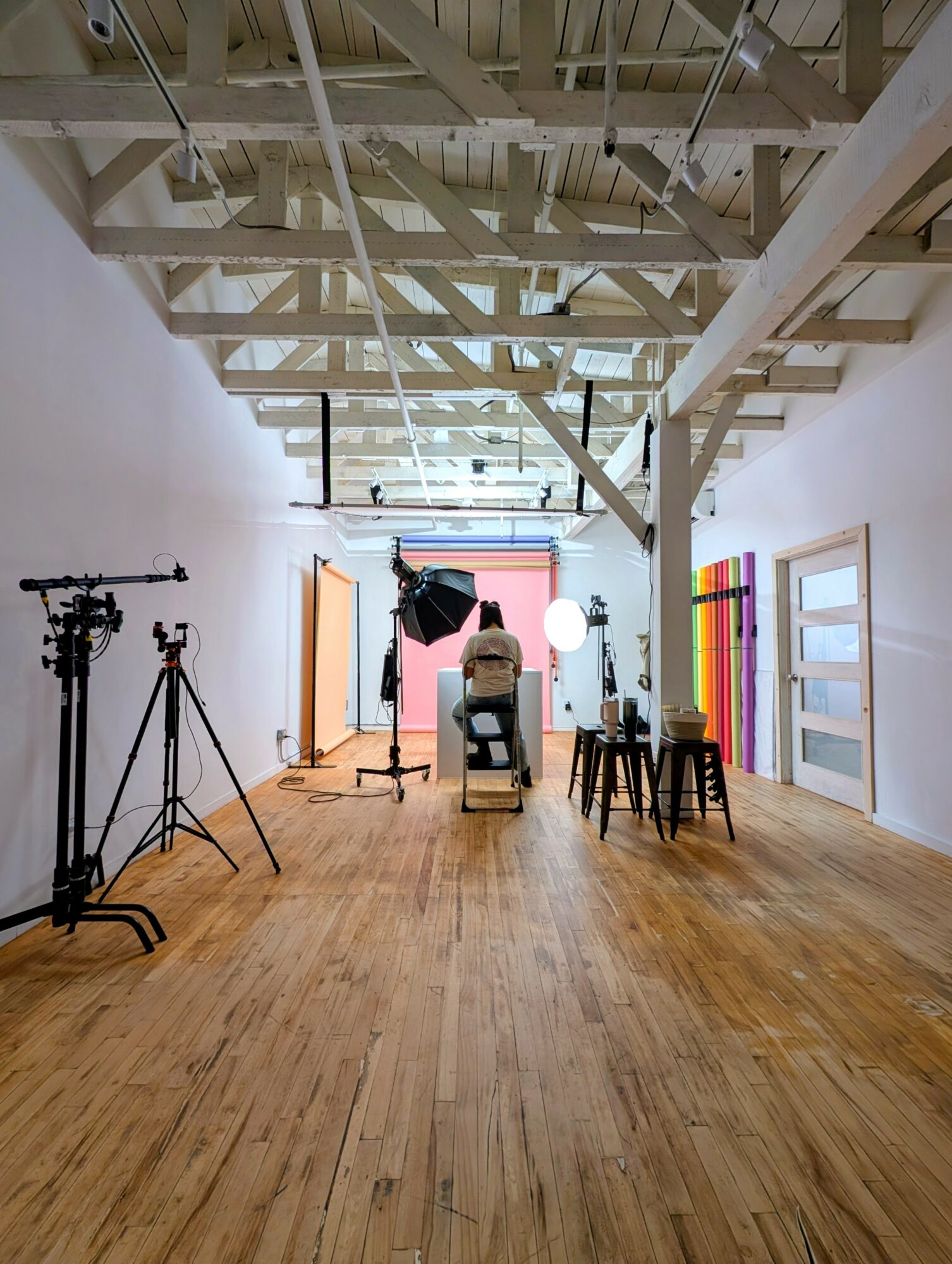 Photography studio with wooden floor, white walls, ceiling beams, and various lighting equipment and chairs.