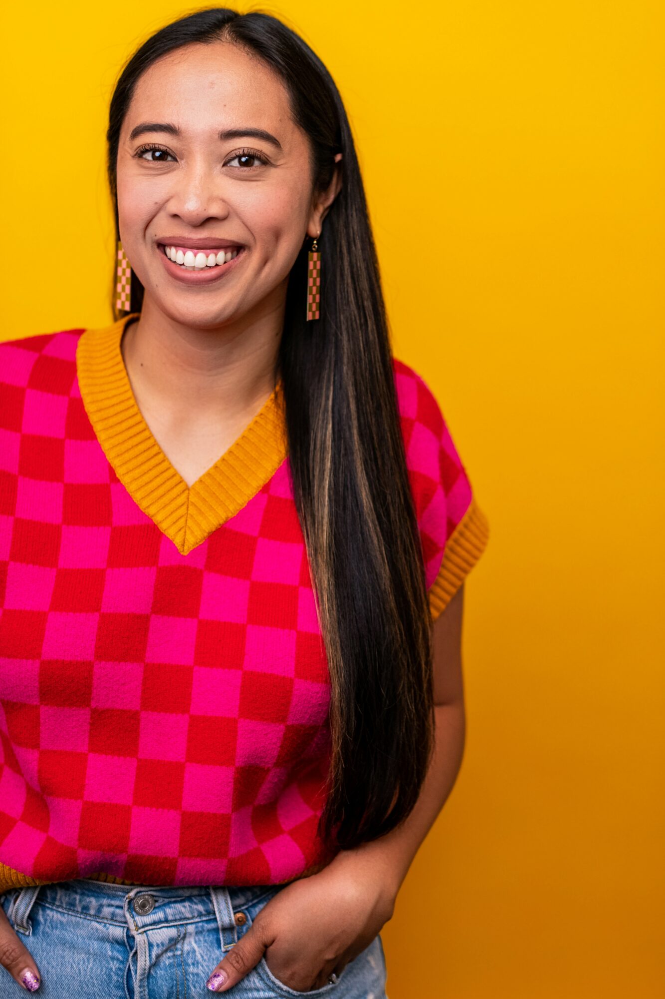 Smiling woman with long dark hair wearing a pink checkered top and earrings, standing against a yellow background.