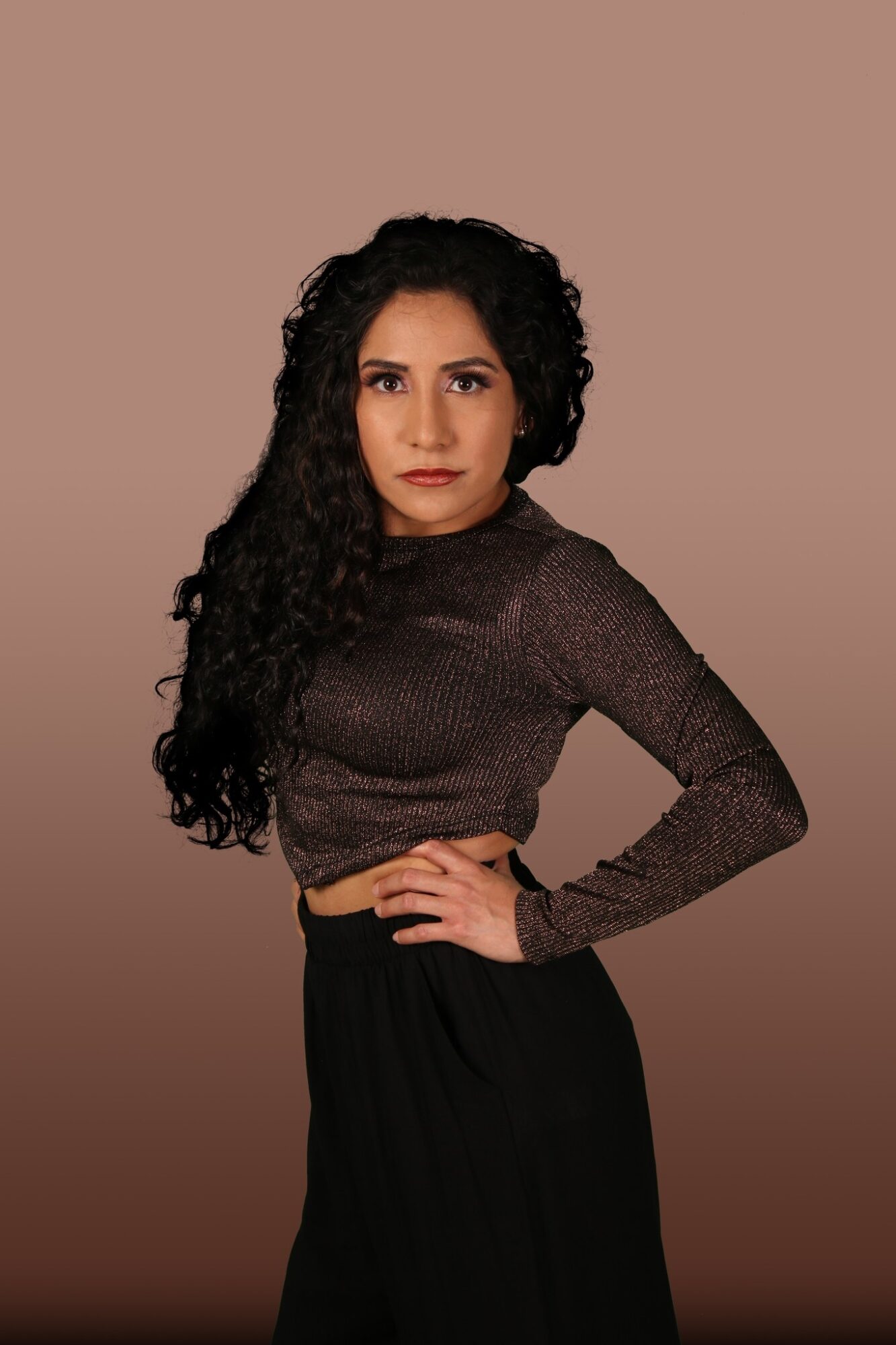Woman with long curly hair wearing a dark long-sleeve top and black skirt, standing with hand on hip against plain background.