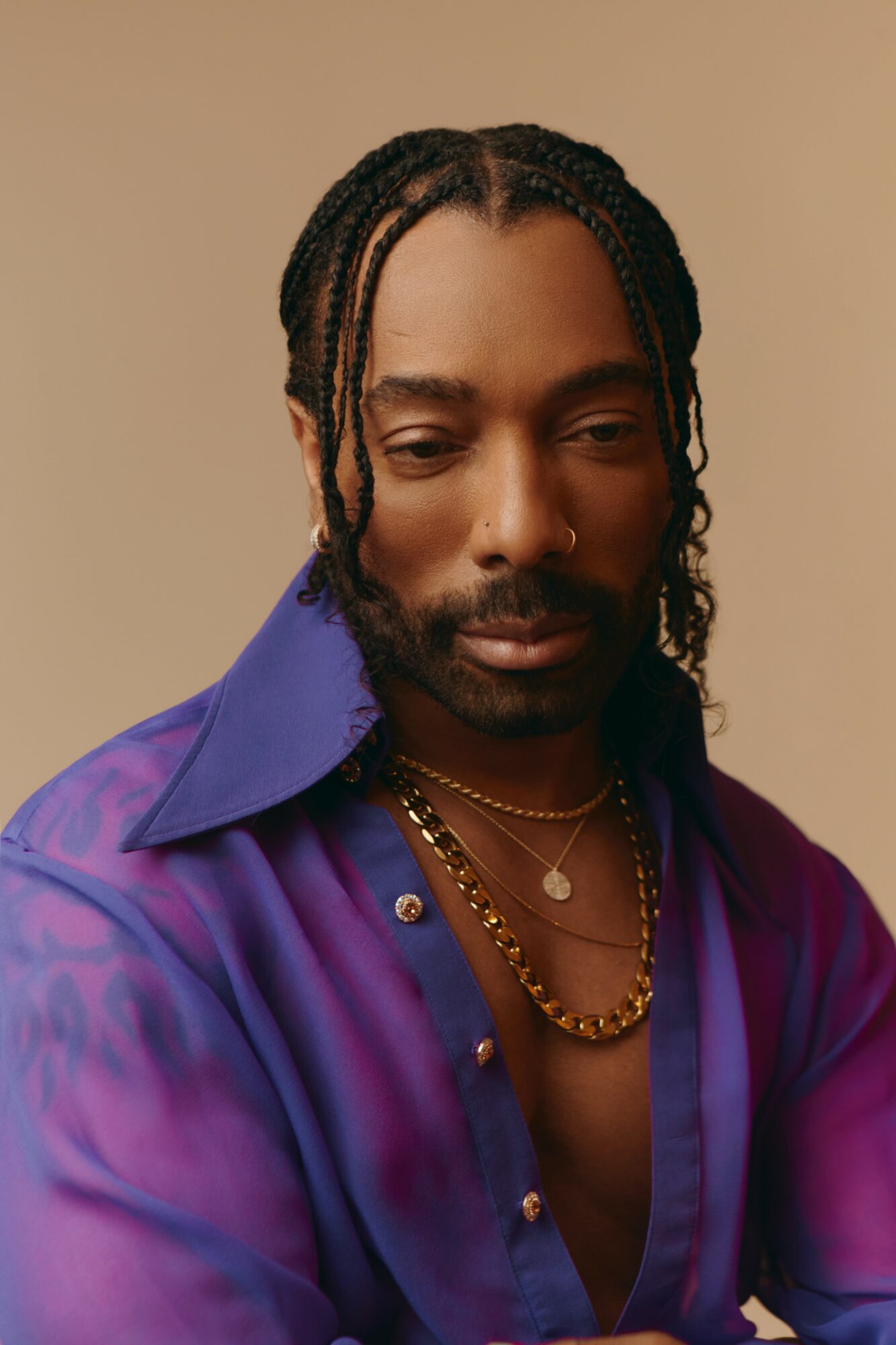 Man with braided hair wearing a purple shirt and gold jewelry, looking at the camera.