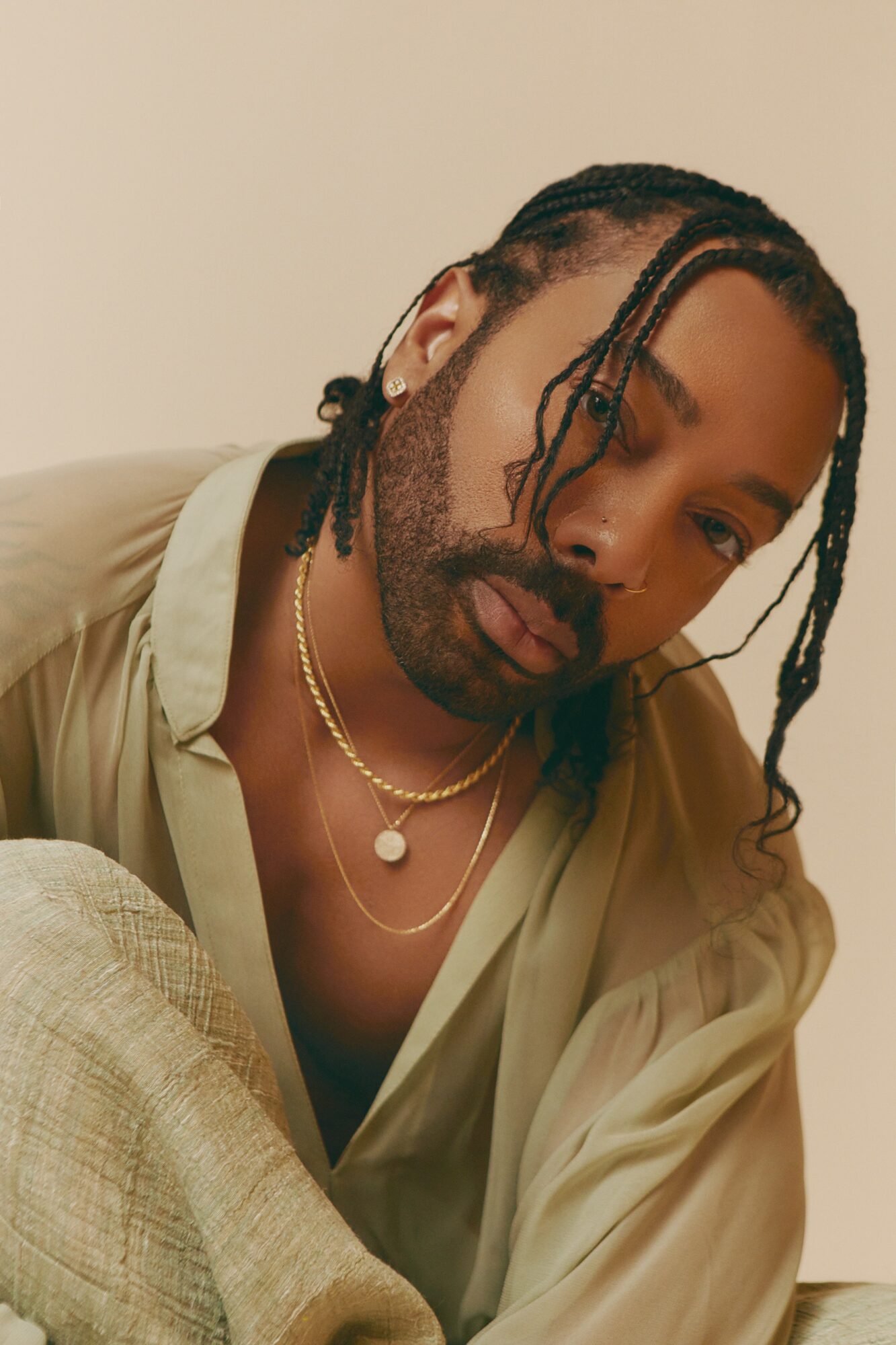 Person with braided hair, wearing layered necklaces and a beige shirt, looking at the camera.