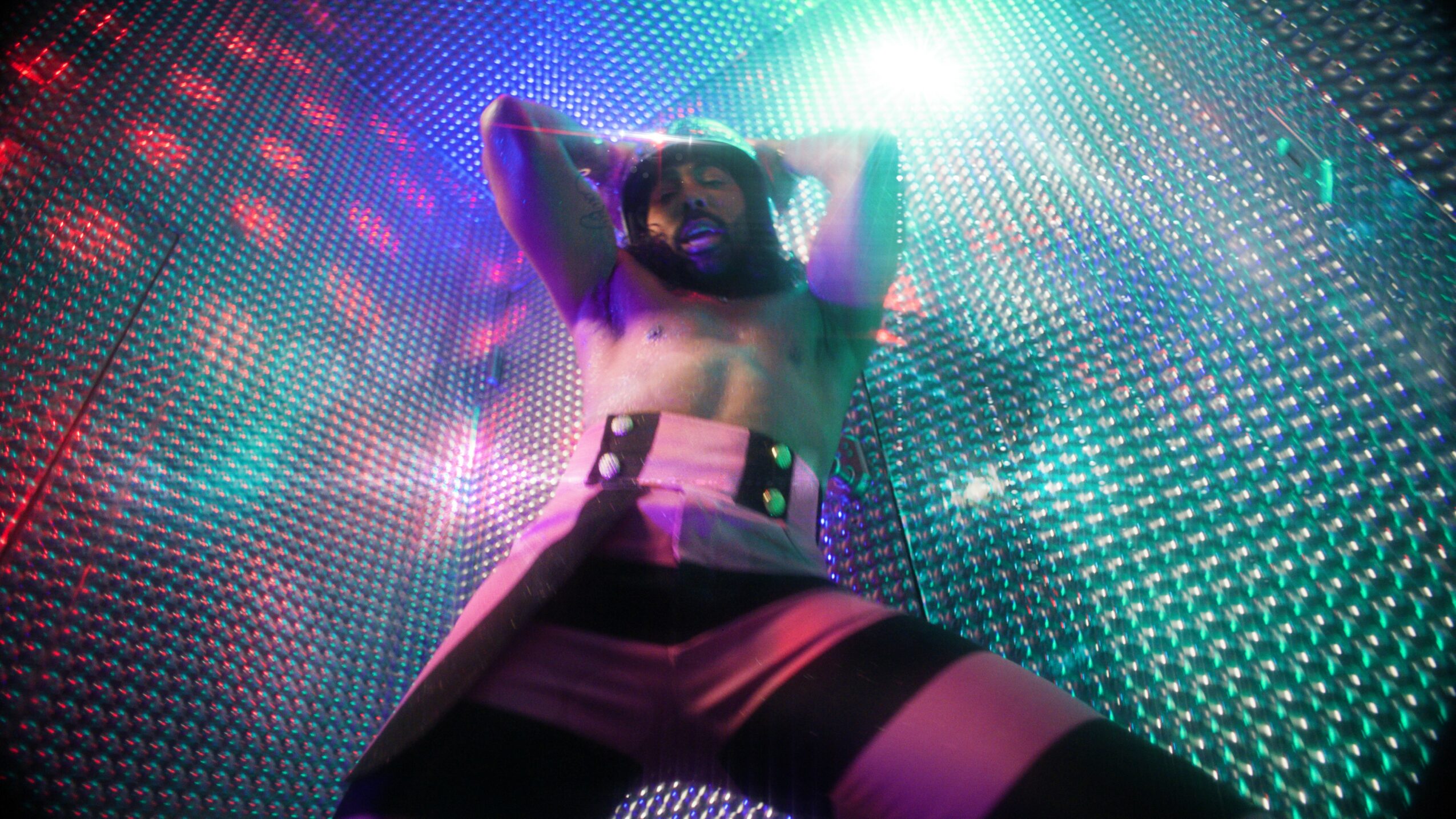 Person with arms raised, standing in front of colorful LED lights and patterns, wearing shorts and a belt.