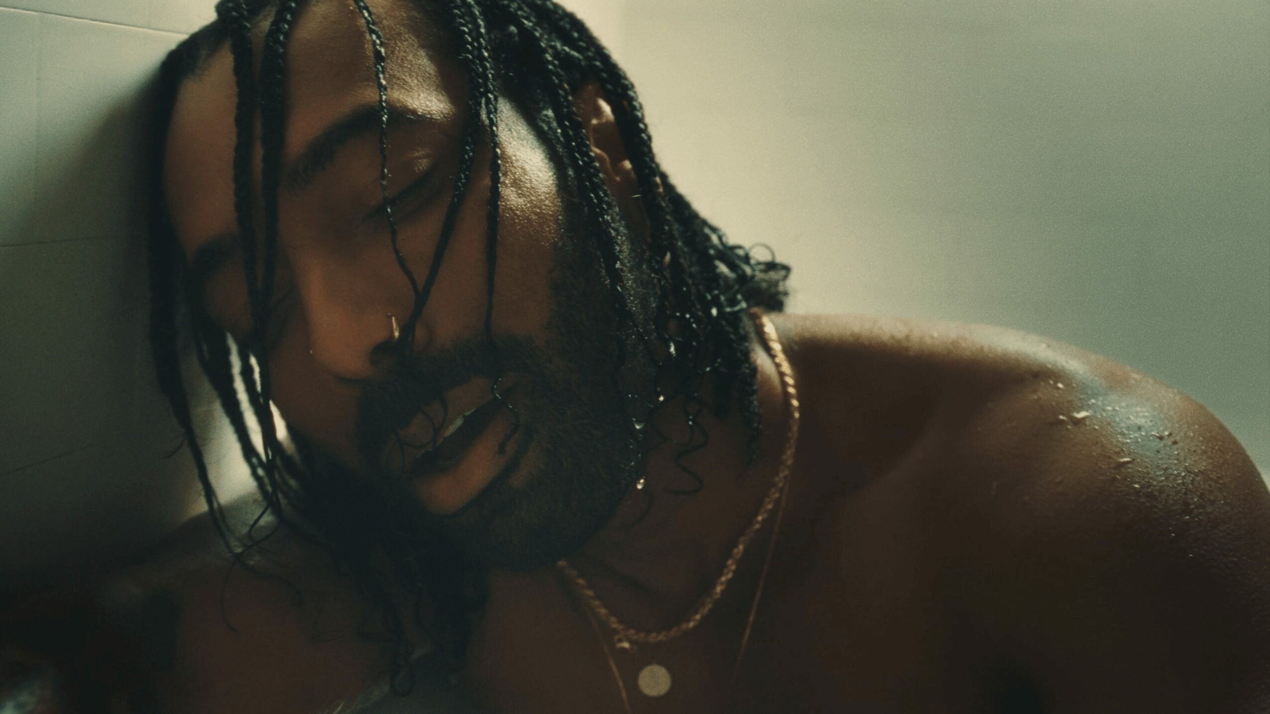 Close-up of a person with braided hair, smiling, with water droplets on skin, wearing necklaces, in a dimly lit setting.
