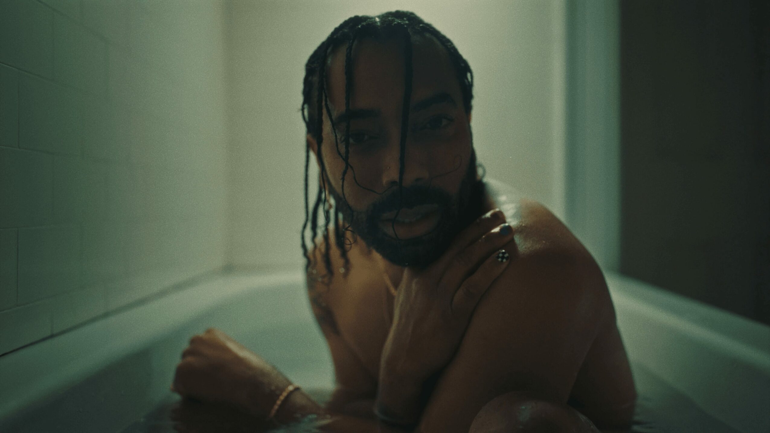 Person with wet hair and beard in a bathtub, looking at the camera, with a neutral expression.