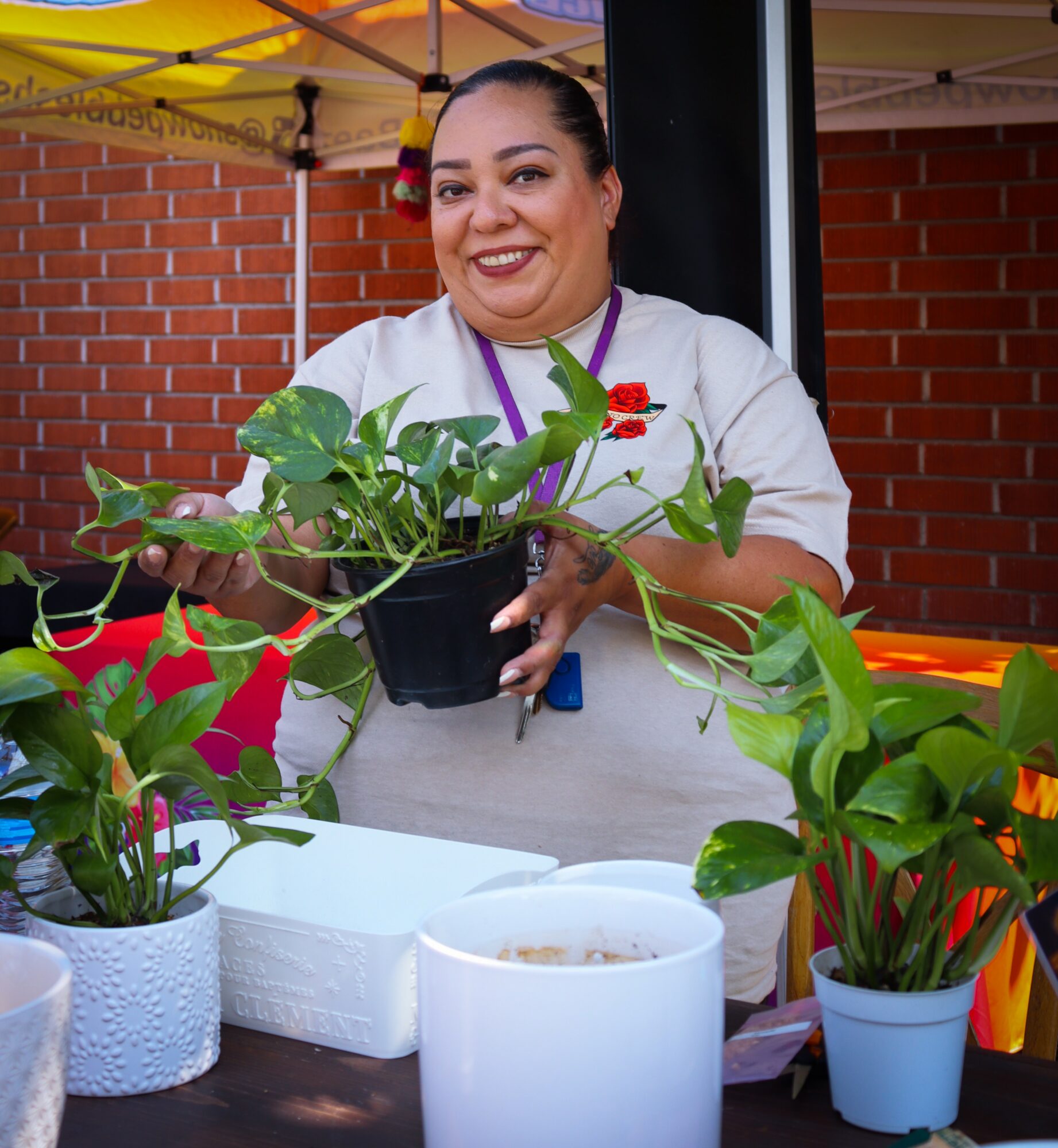 Smiling woman holding a potted plant at an outdoor market stall with colorful plants and brick wall background.