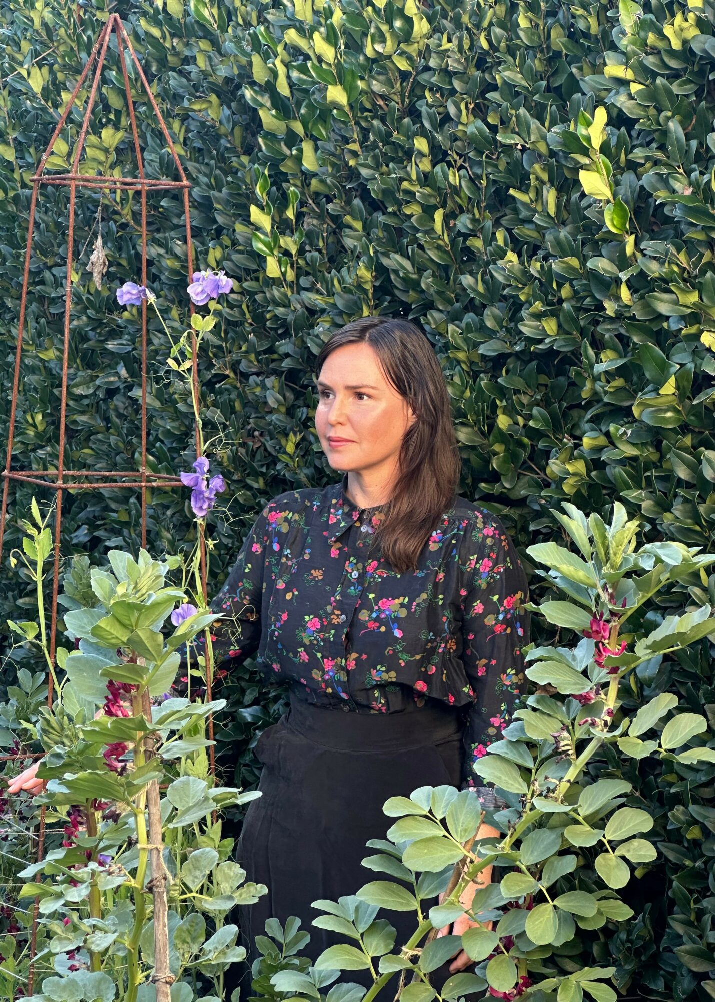 Woman with shoulder-length hair standing among green plants and purple flowers outdoors.