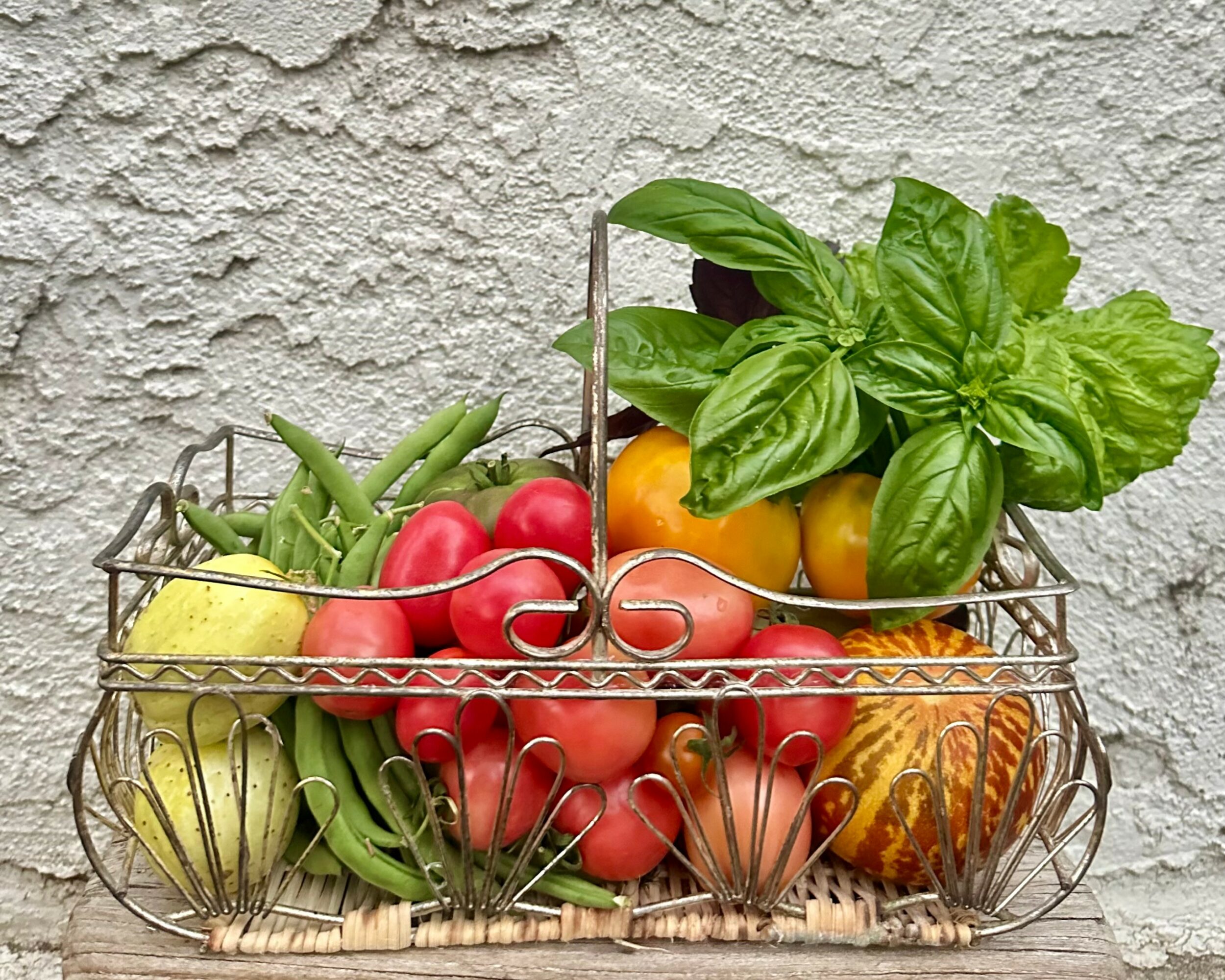 Wire basket filled with tomatoes, apples, bananas, lemon, and basil leaves against a textured wall.