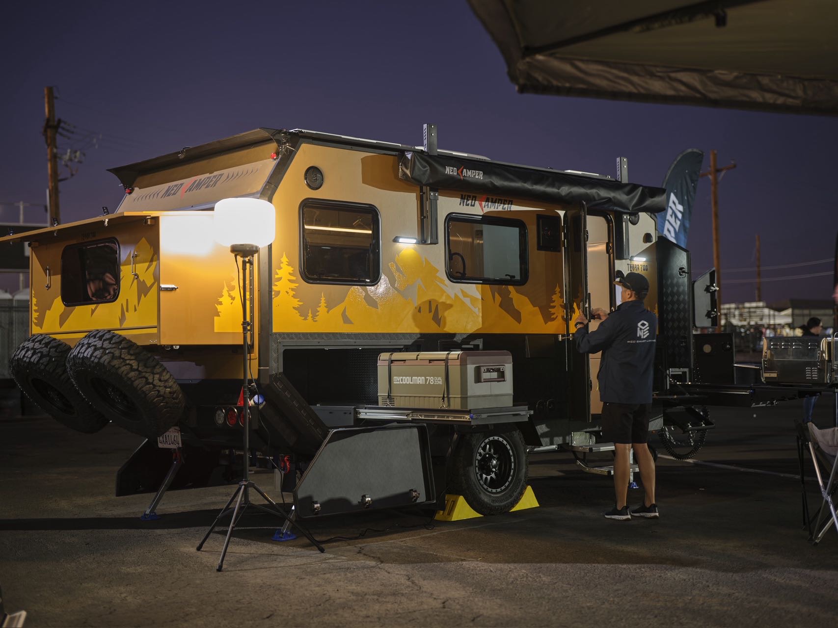 Yellow and black camper trailer with a person standing nearby at dusk, outdoor setting with equipment around.