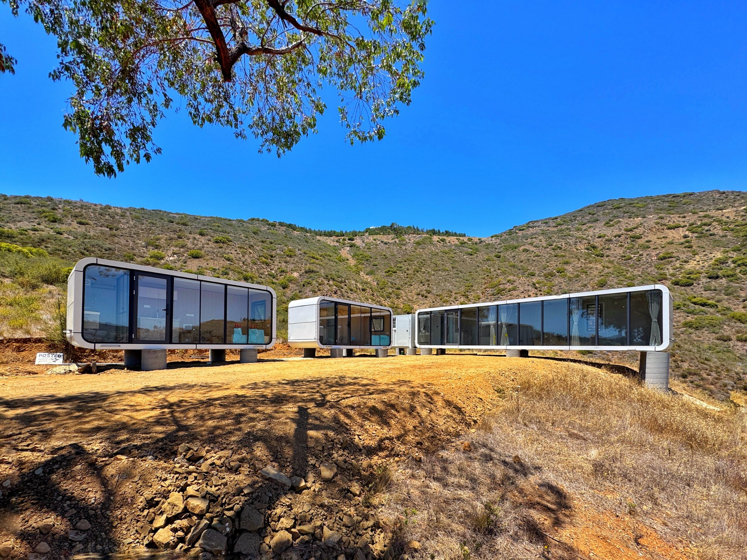 Three modern glass buildings on a dirt ground with hills and a tree in the background under a clear blue sky.