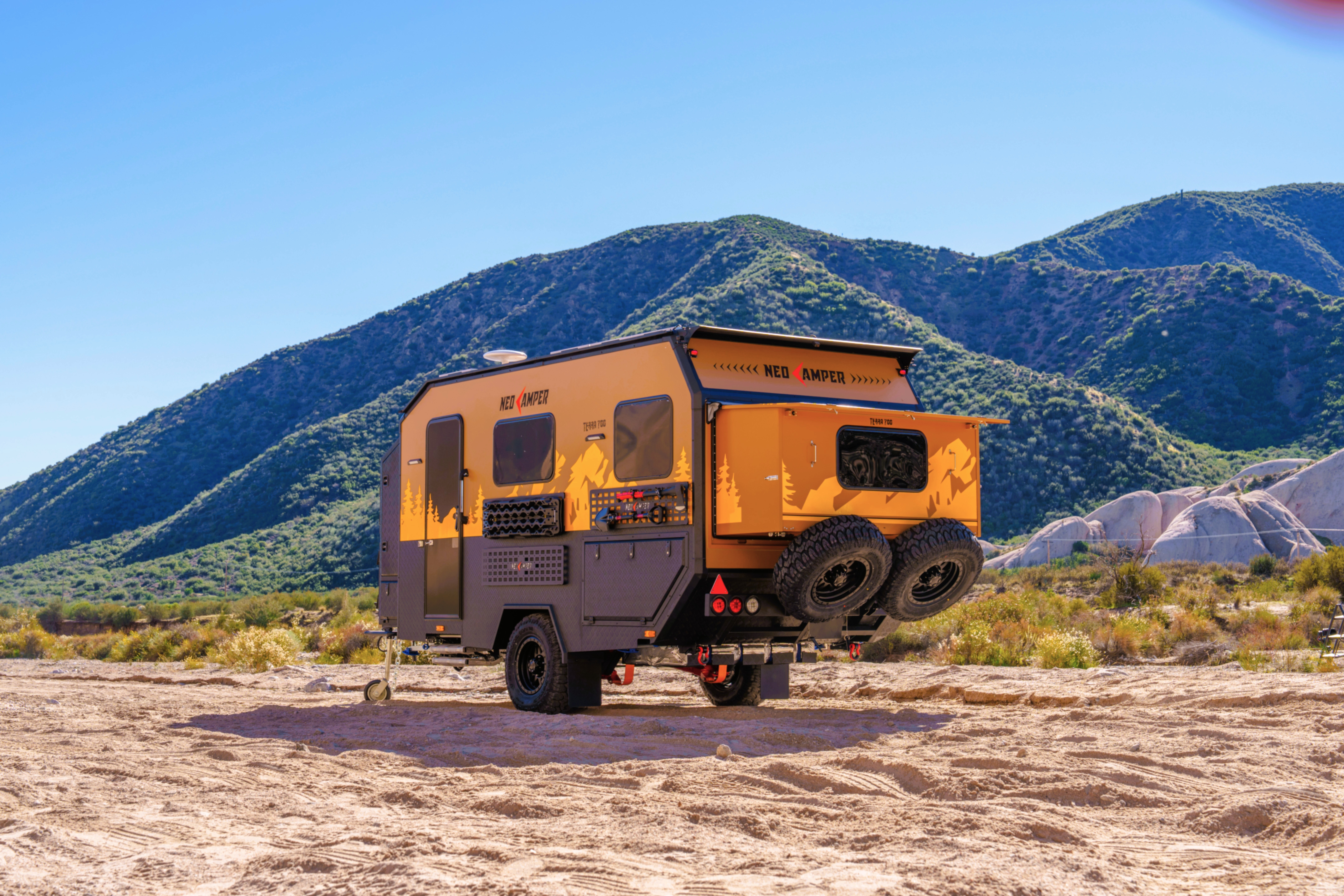 Orange and black camper trailer parked on a dirt area with mountains and blue sky in background.