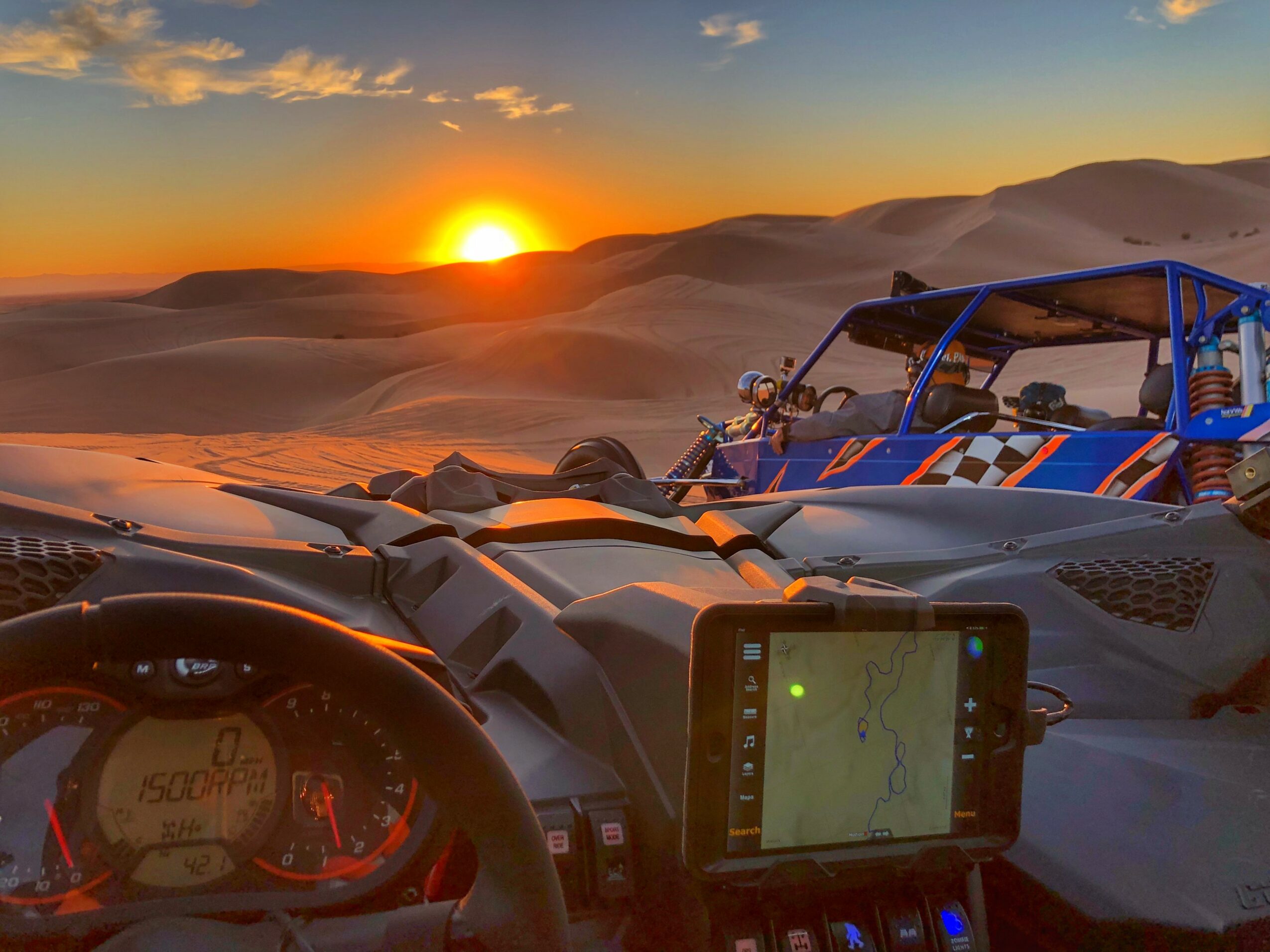 Off-road vehicles parked on sand dunes during sunset with a GPS device and dashboard visible.