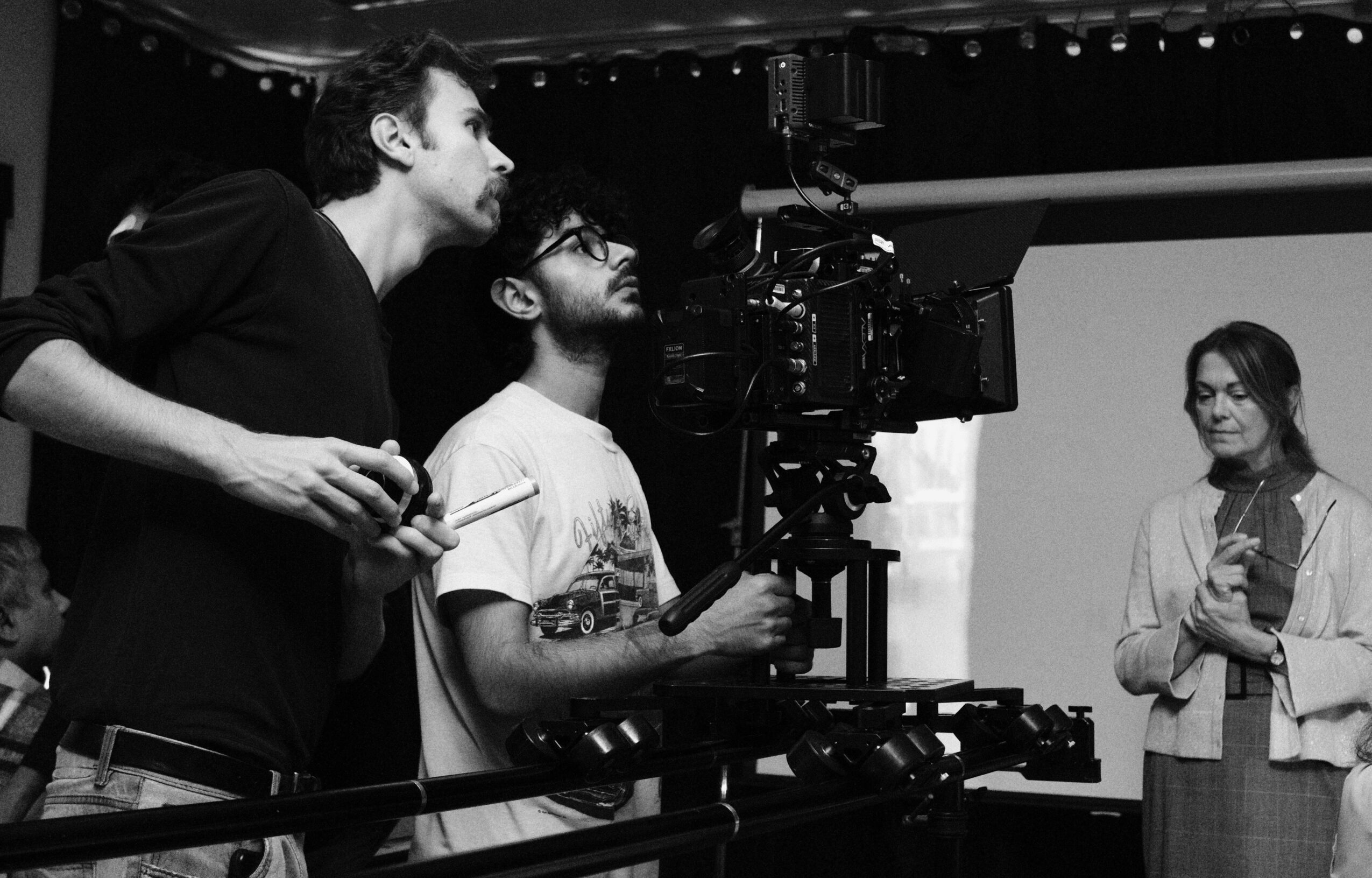 Two men operate a camera on a film set, with a woman standing nearby in front of a screen.