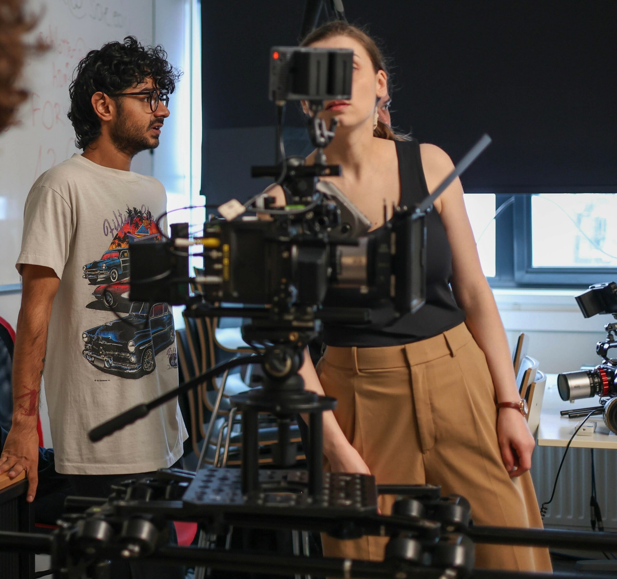 Two people in a room with camera equipment, one woman operating a camera, one man observing.