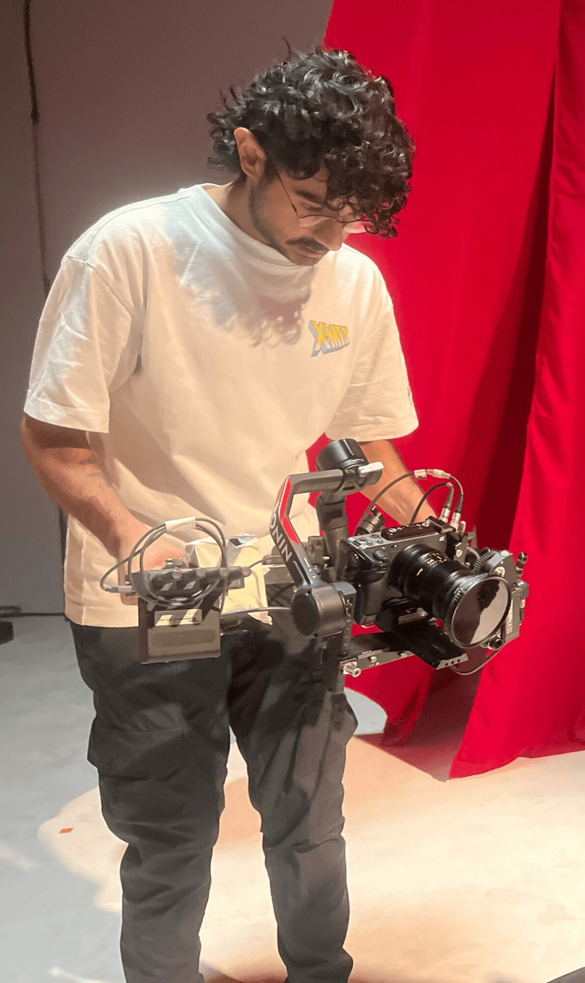 Person with curly hair wearing a white T-shirt and dark pants, operating a camera with a red and black rig.