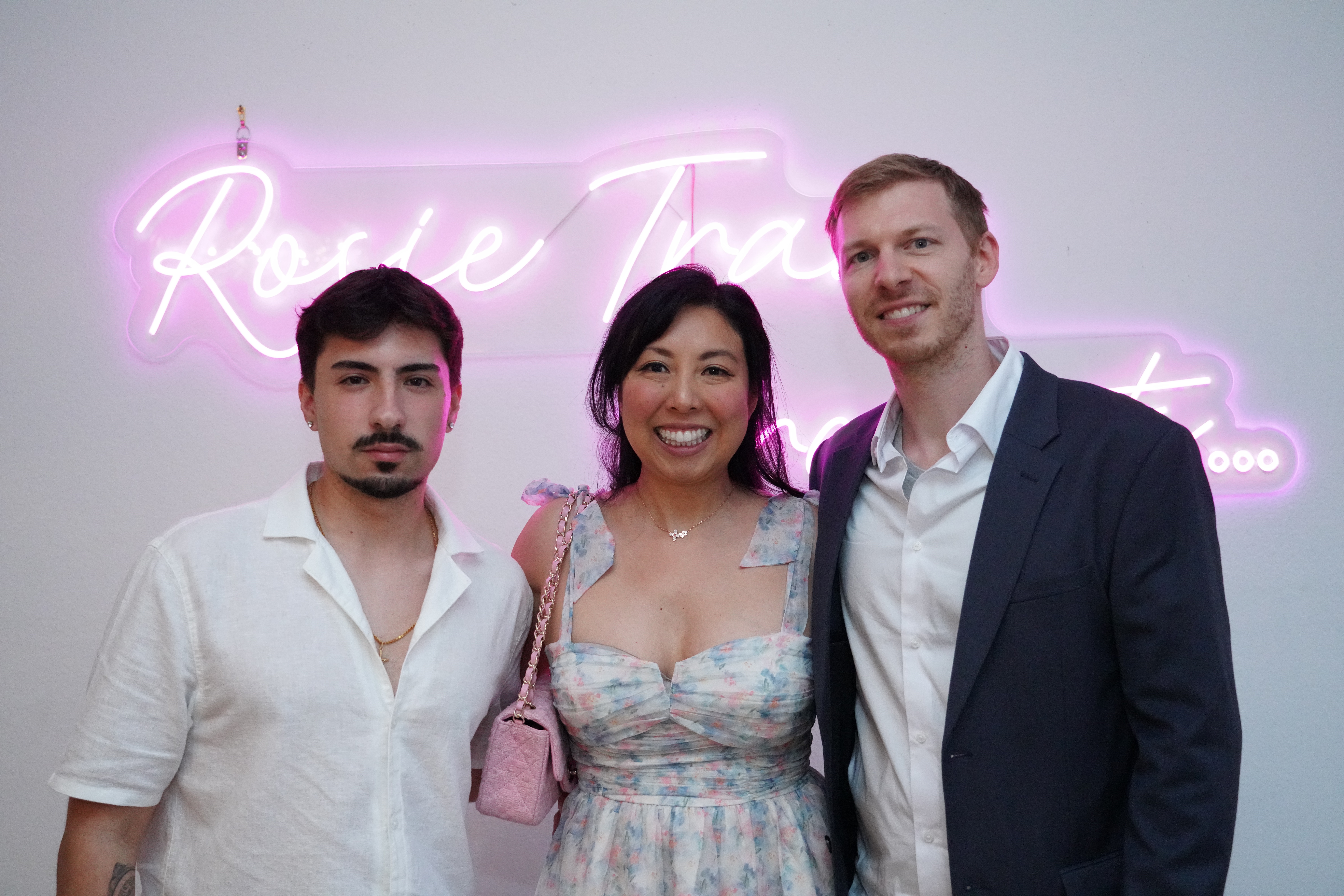 Three people standing together in front of a neon sign, smiling, in a well-lit room.