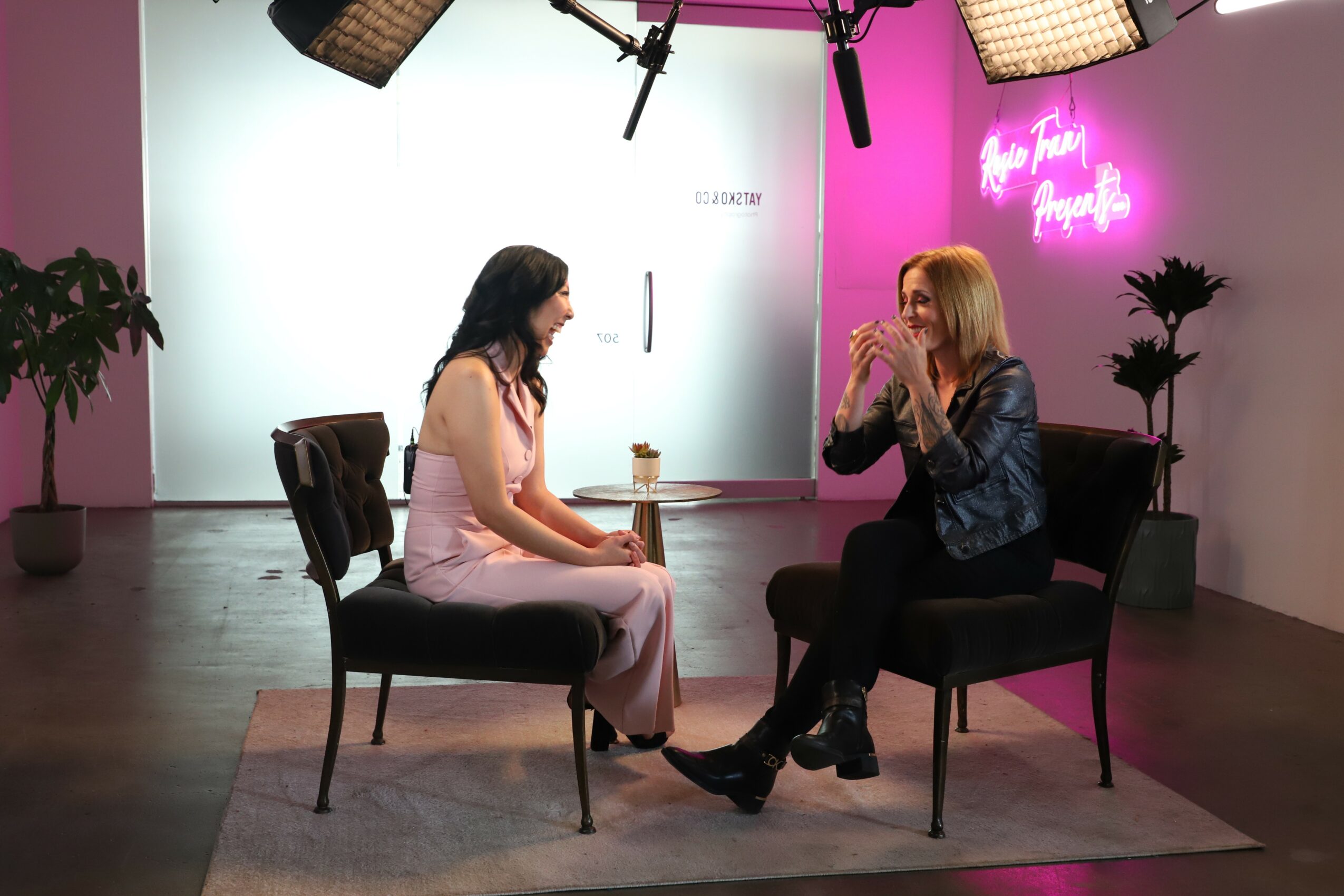 Two women sit on chairs in a studio with lighting equipment, plants, and a pink neon sign in the background.