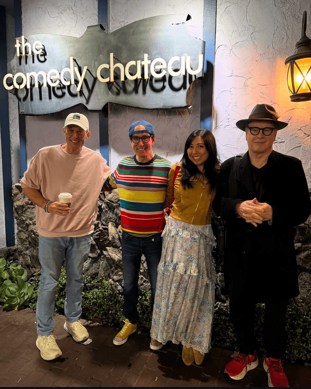 Four people standing together in front of a comedy club sign, smiling, with plants and a wall lamp nearby.