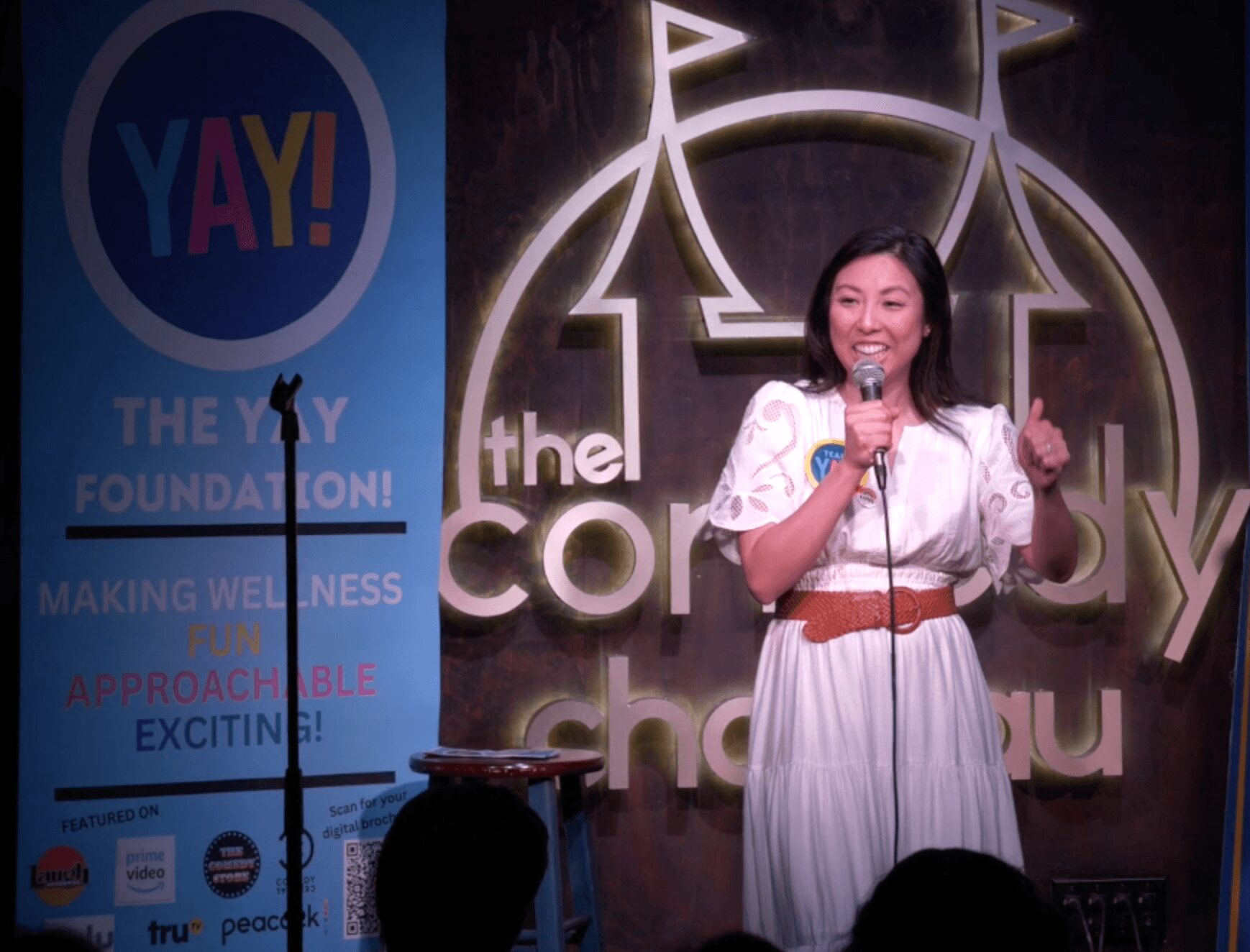 Woman speaking on stage with a microphone, smiling, wearing a white dress with a brown belt, in front of a sign.