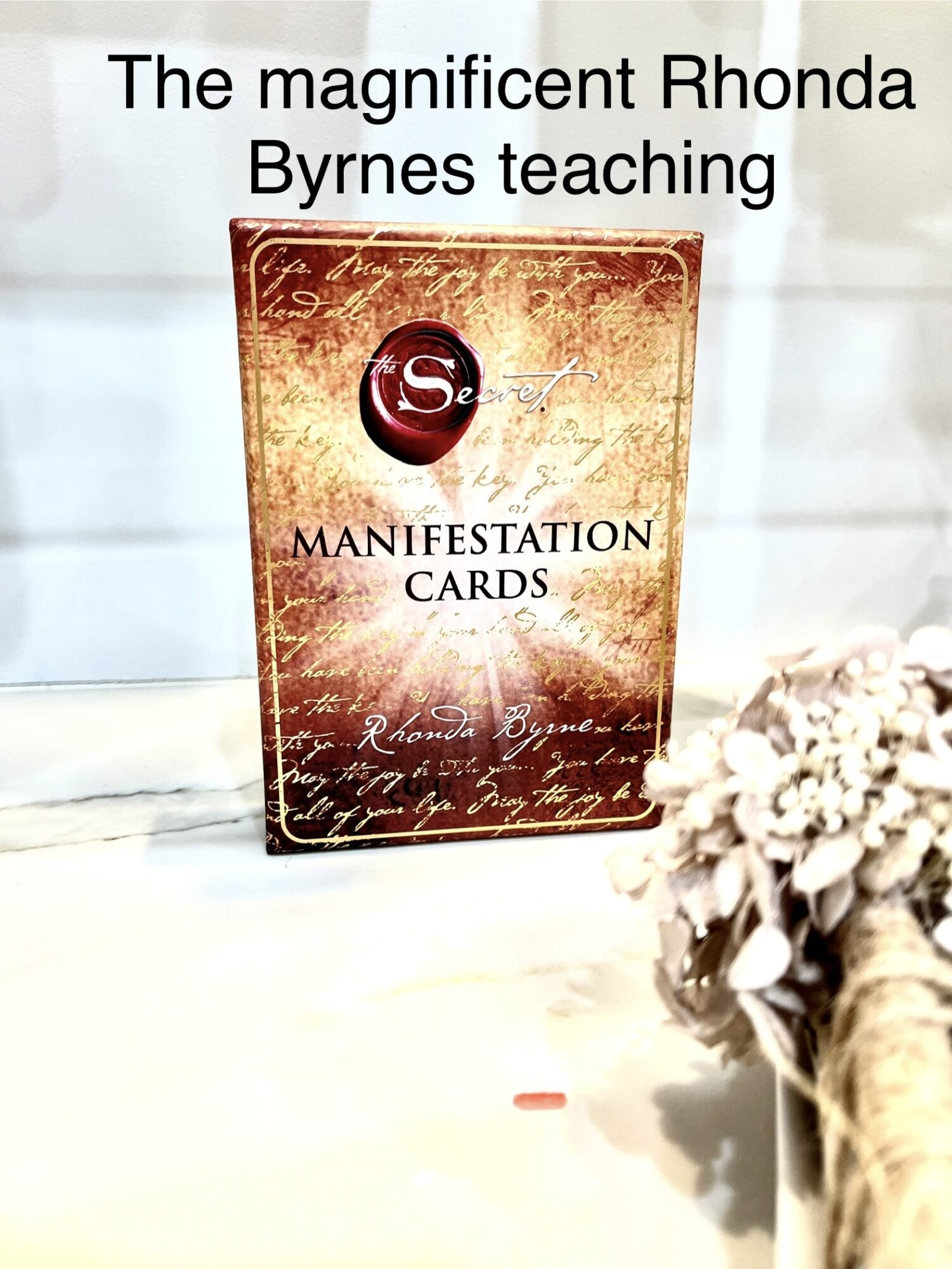 A card deck labeled 'Manifestation Cards' stands upright with handwritten script background, next to a bouquet of white flowers.