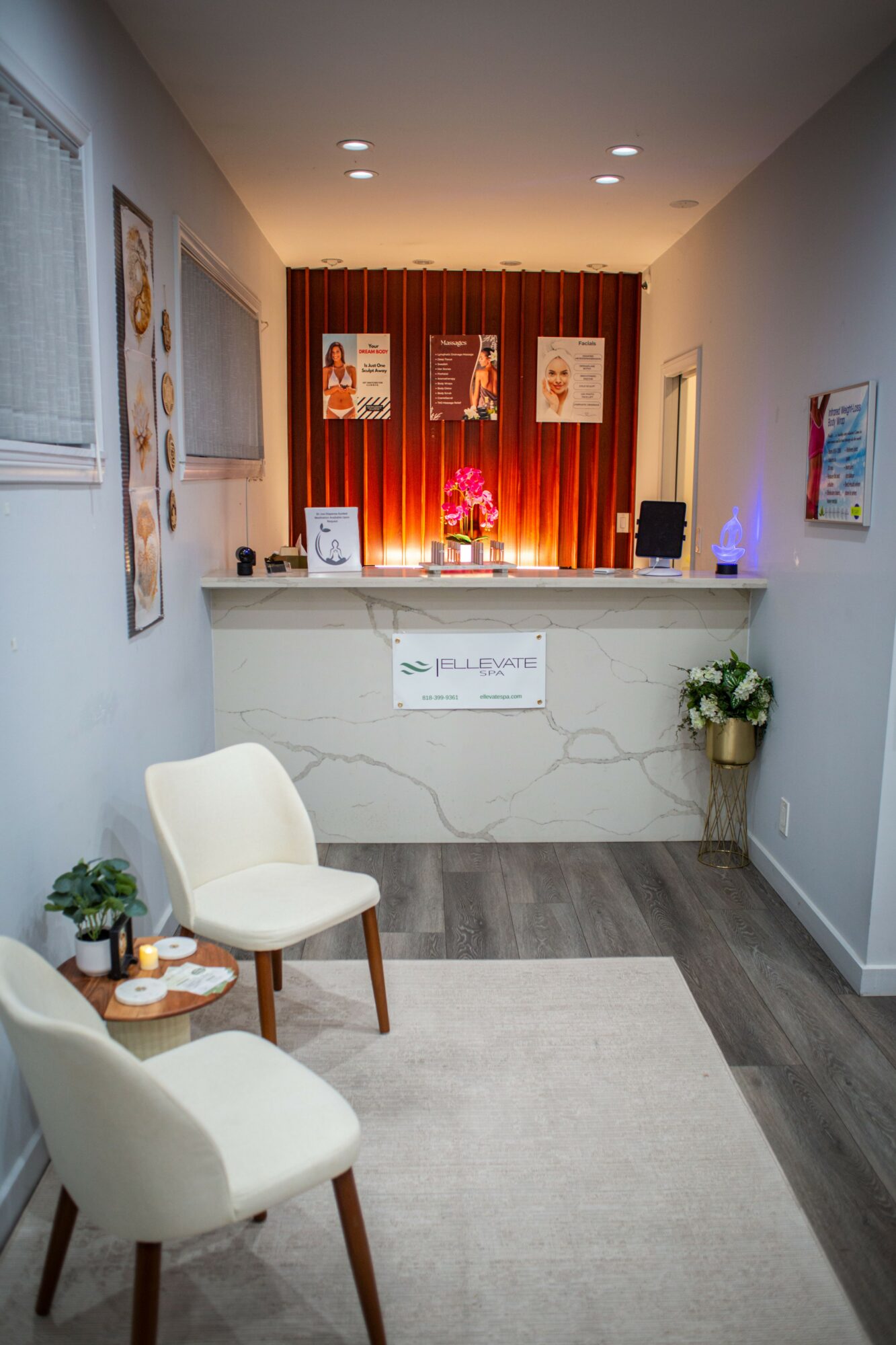 Reception area with white chairs, a small table, and a marble counter. Decor includes framed artwork and a potted plant.