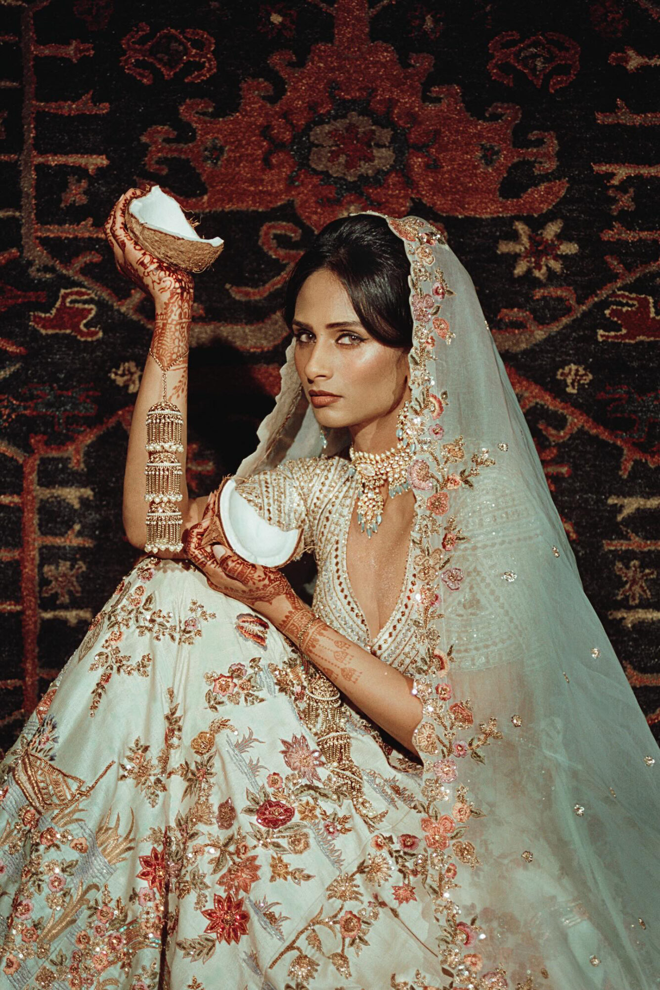 Woman in traditional attire holding a white object, sitting on a patterned rug with intricate designs.