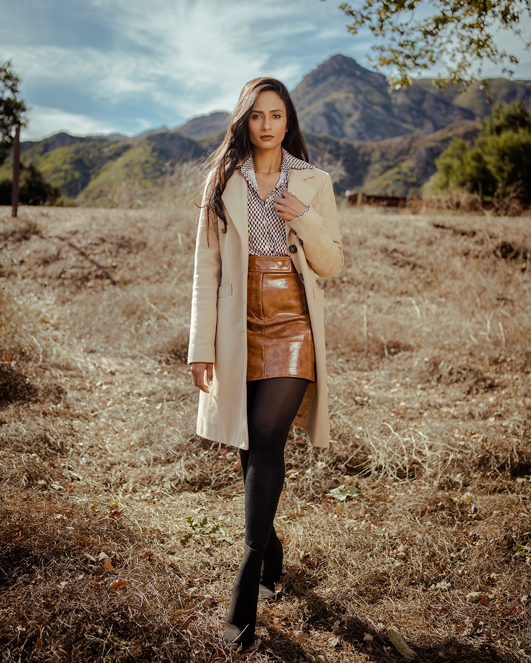 Young woman standing outdoors in a field with mountains and trees in background, wearing a coat, dress, and tights.